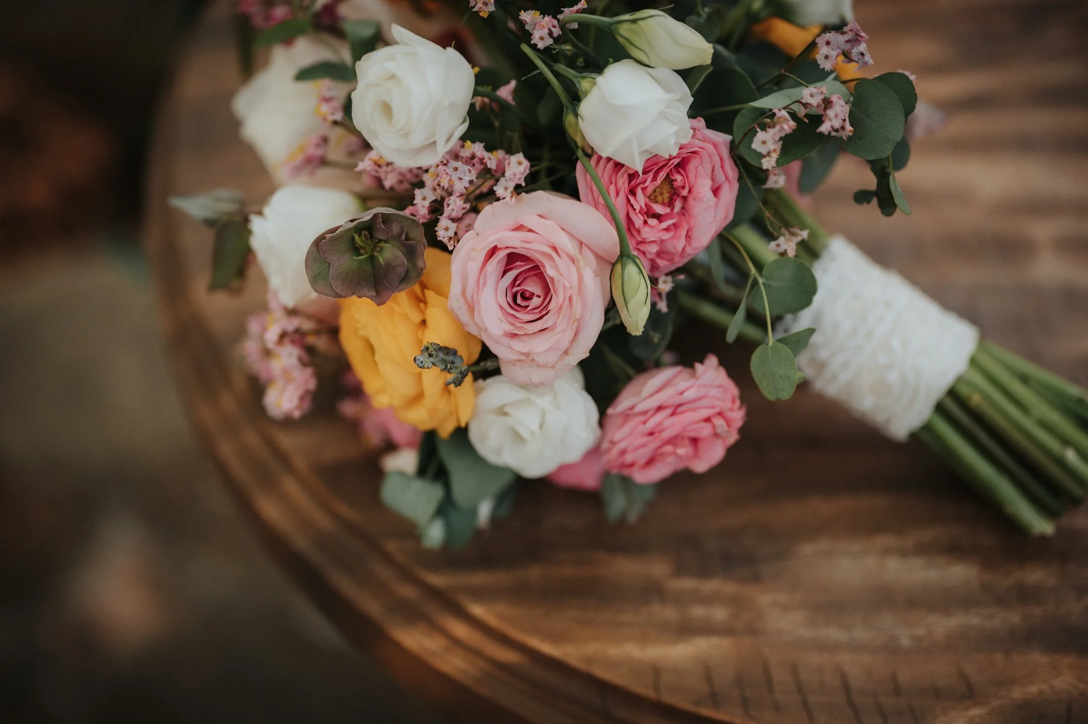 A bouquet of various colored flowers, including pink, white, and yellow roses, resting on a wooden surface, with a lace-wrapped stem.
