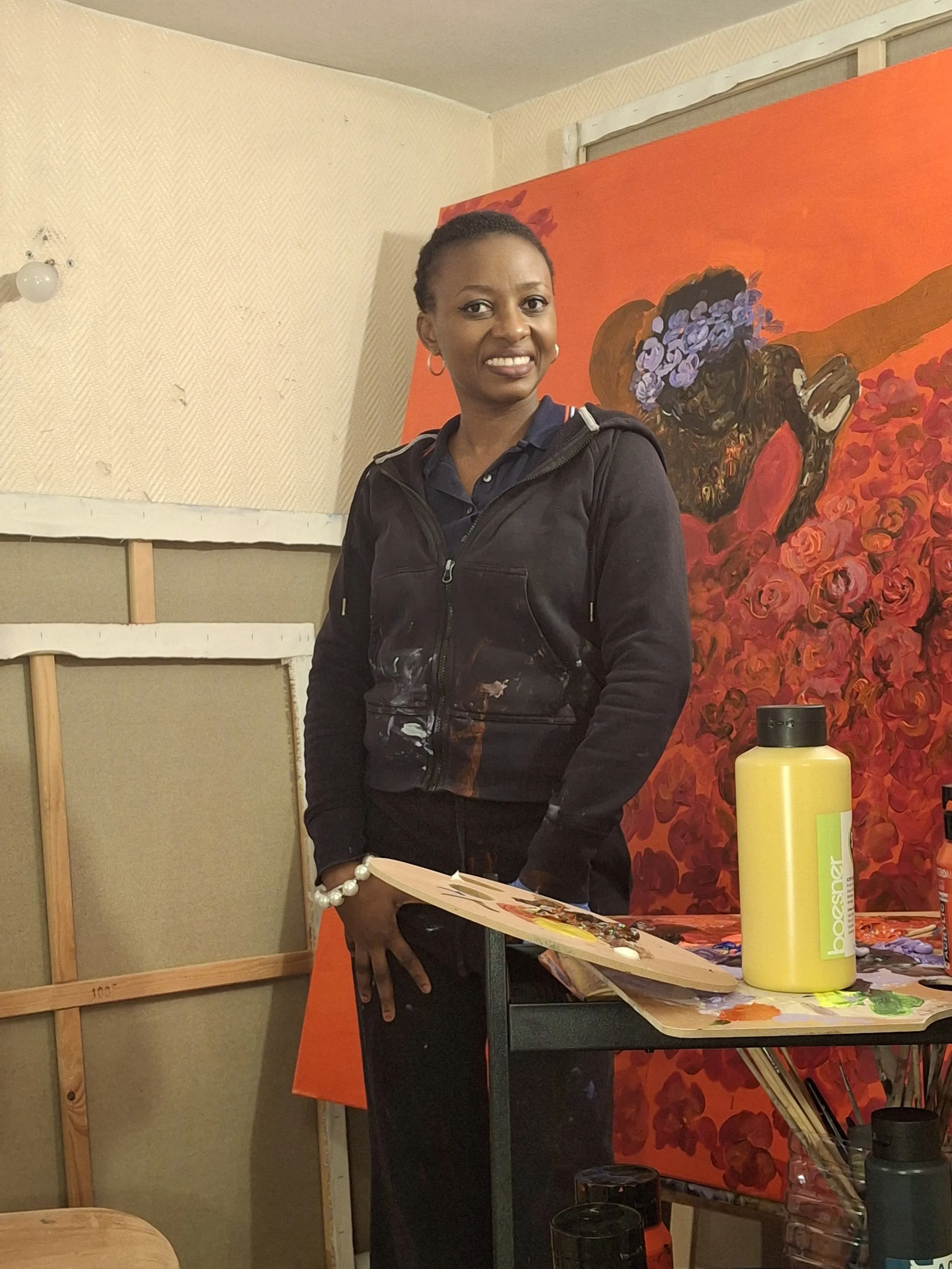 Une femme souriante en veste noire, debout devant une peinture de fleurs violettes et roses sur fond orange, dans un atelier d'art.