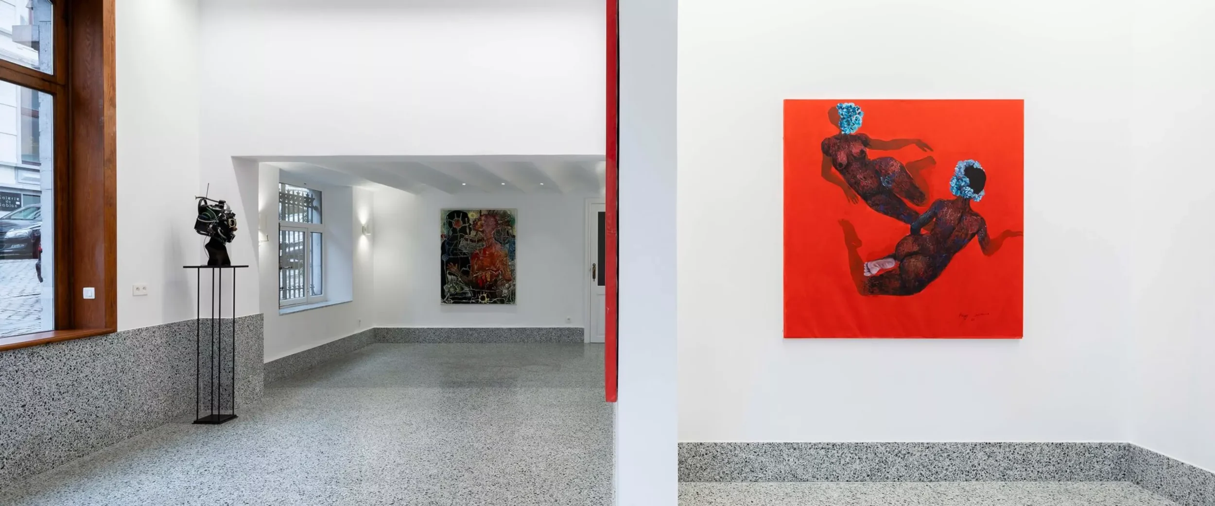 Exposition d'art avec sculptures et peintures dans une galerie, deux figures féminines peintes en rouge avec des coiffures bleues sur un mur blanc.