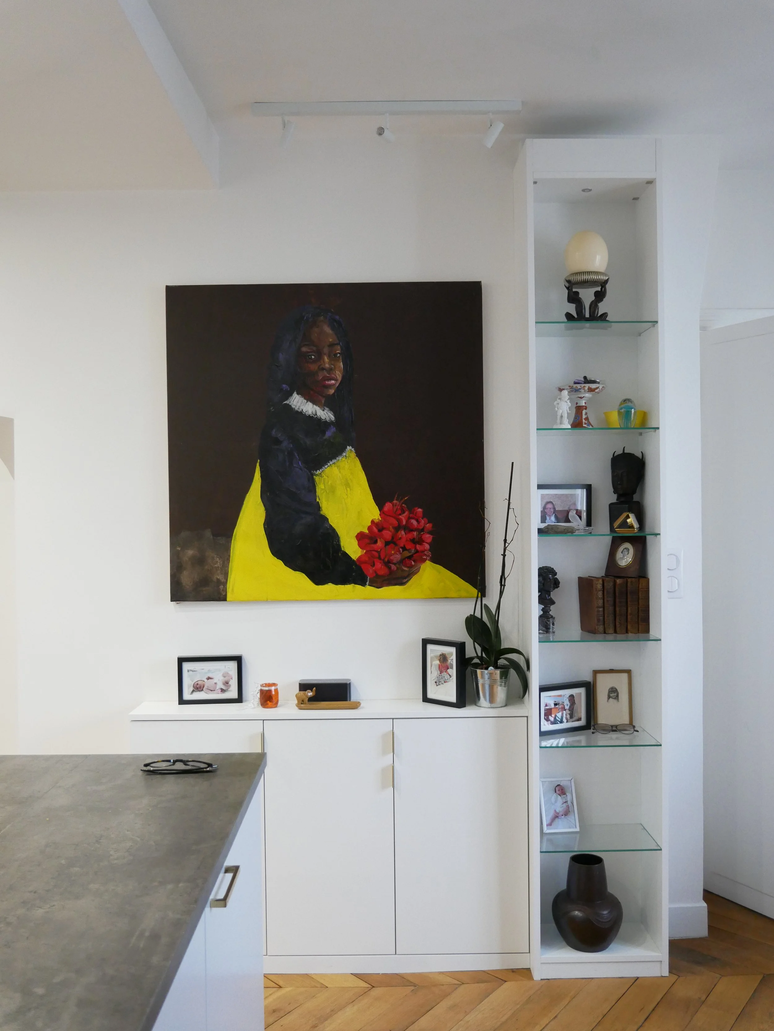Un mur blanc avec une grande peinture représentant une femme noire portant une robe jaune et une veste noire, tenant un bouquet de fleurs rouges. À droite, une étagère intégrée avec des décorations, des photos de famille et des livres.