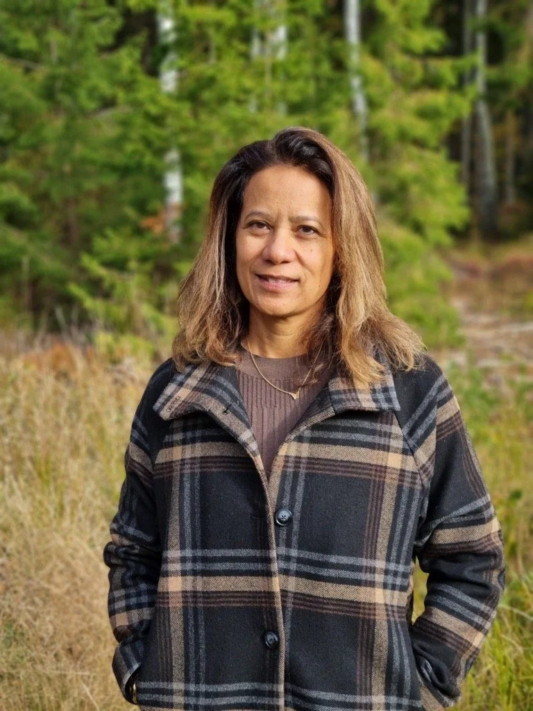 A woman standing outdoors in a forested area during daytime, wearing a plaid coat and looking at the camera.