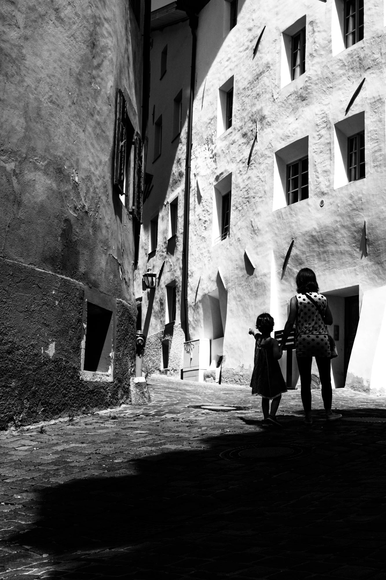 Schwarz-Weiß-Foto einer engen, kurvigen Gasse mit alten Gebäuden. Zwei Kinder stehen im Schatten und scheinen miteinander zu sprechen oder zu spielen.