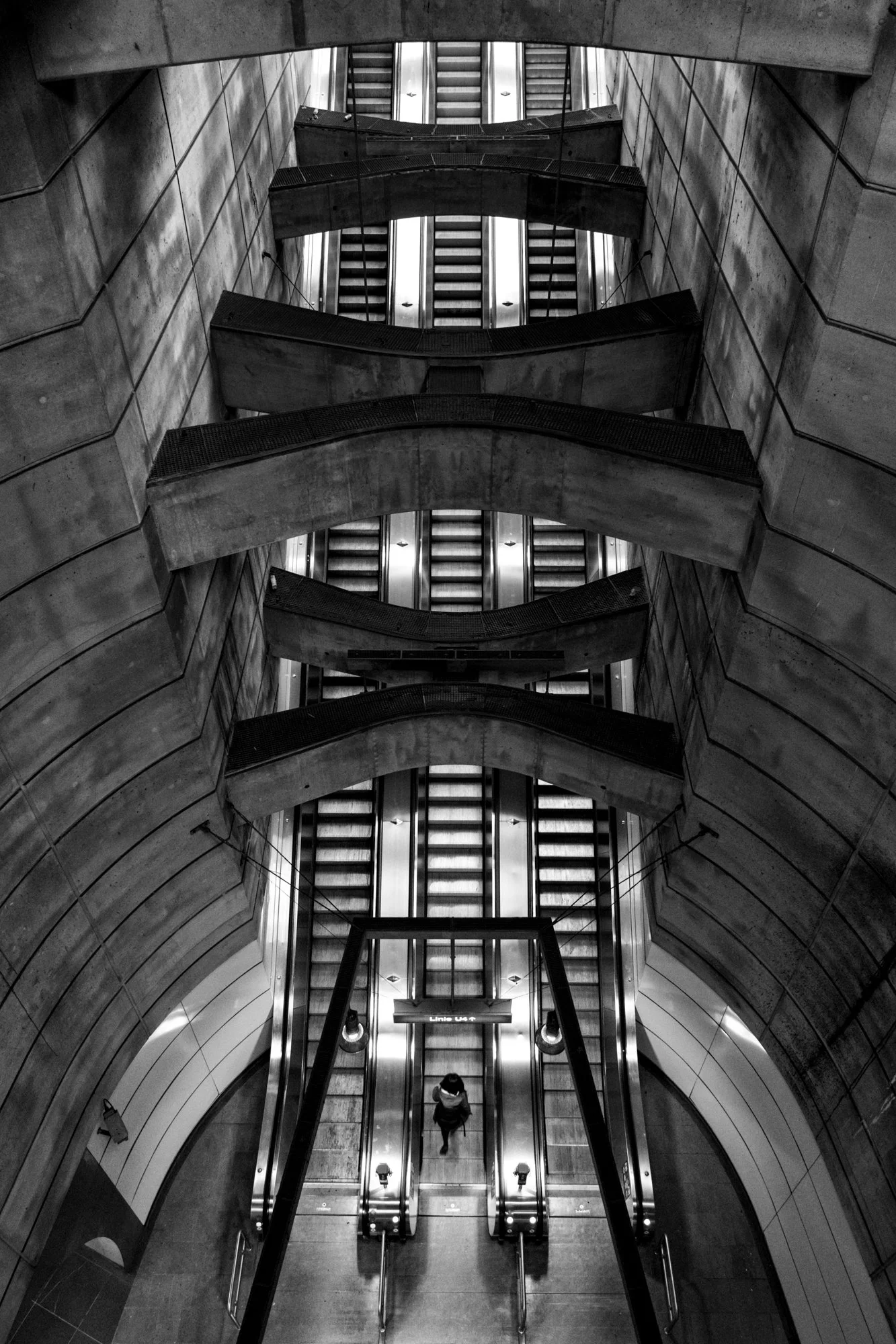 Mehrstöckige gut beleuchtete Rolltreppe in einem modernen, grauen, geschwungenen Gebäude, mit einer einzelnen Person, die nach oben fährt.