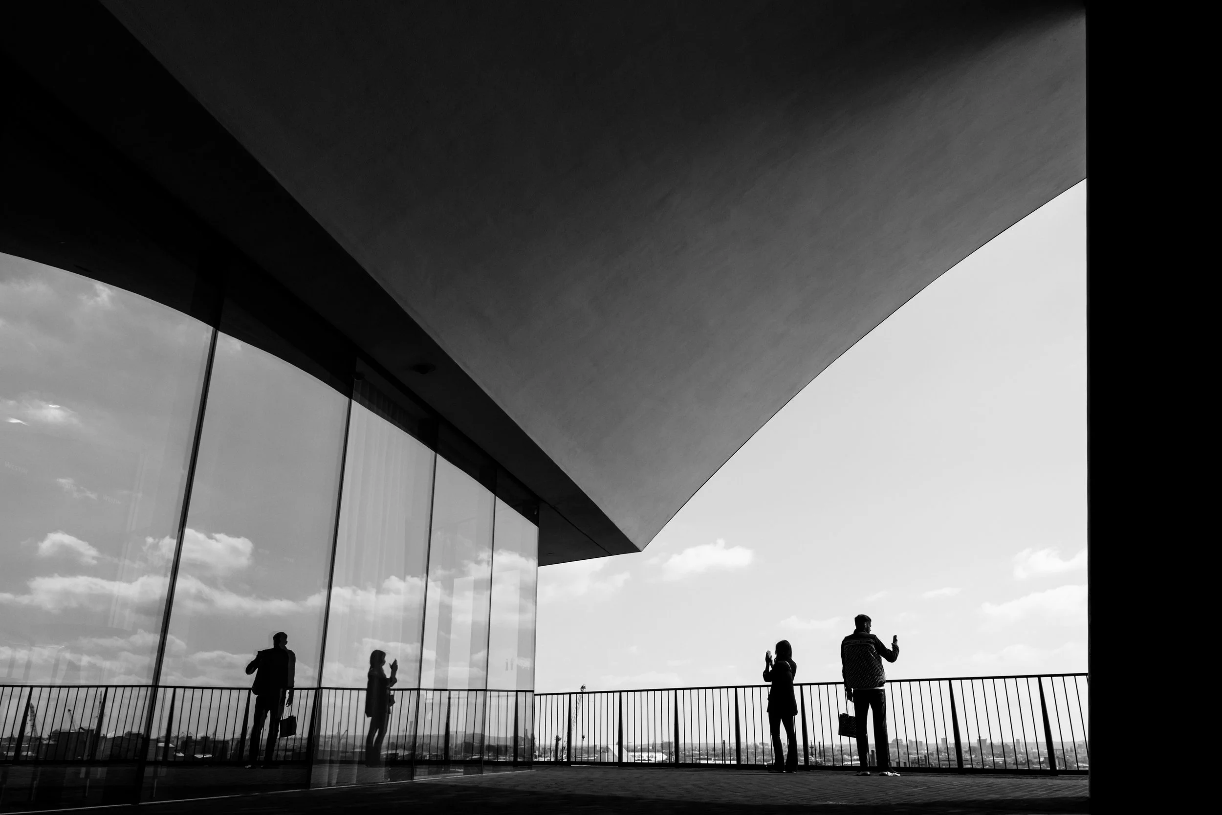 Silhouetten von fünf Menschen, die auf einer Balkonbrüstung vor einem modernen Gebäude mit großen Glasfenstern stehen, während sie Handys benutzen, bei Sonnenlicht mit Himmel und Wolken im Hintergrund.