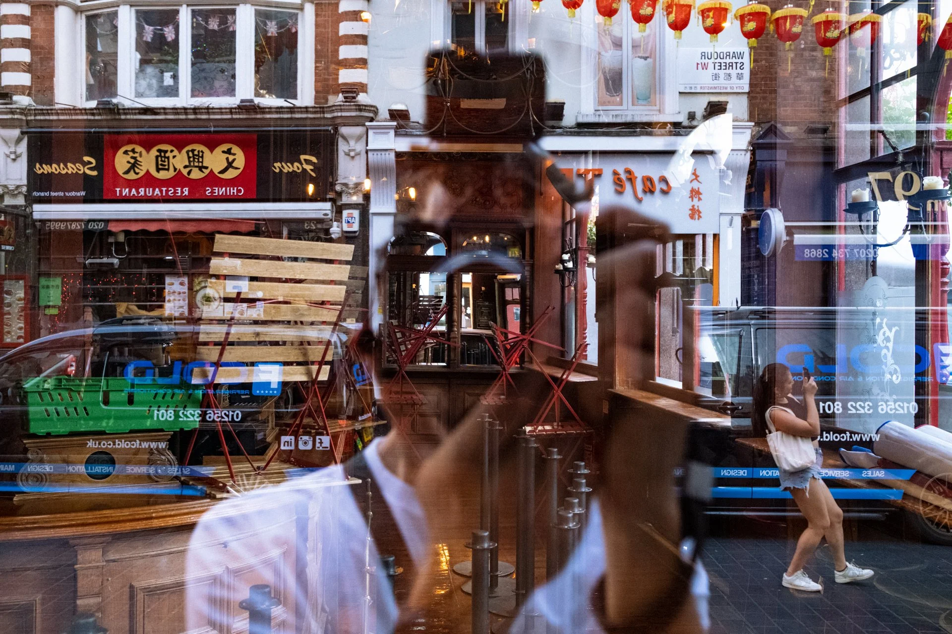Reflexion eines Fußgängers und einer jungen Frau im Schaufenster eines chinesischen Restaurants in einer belebten Straße, mit sichtbaren chinesischen Schriftzeichen und roten Laternen.