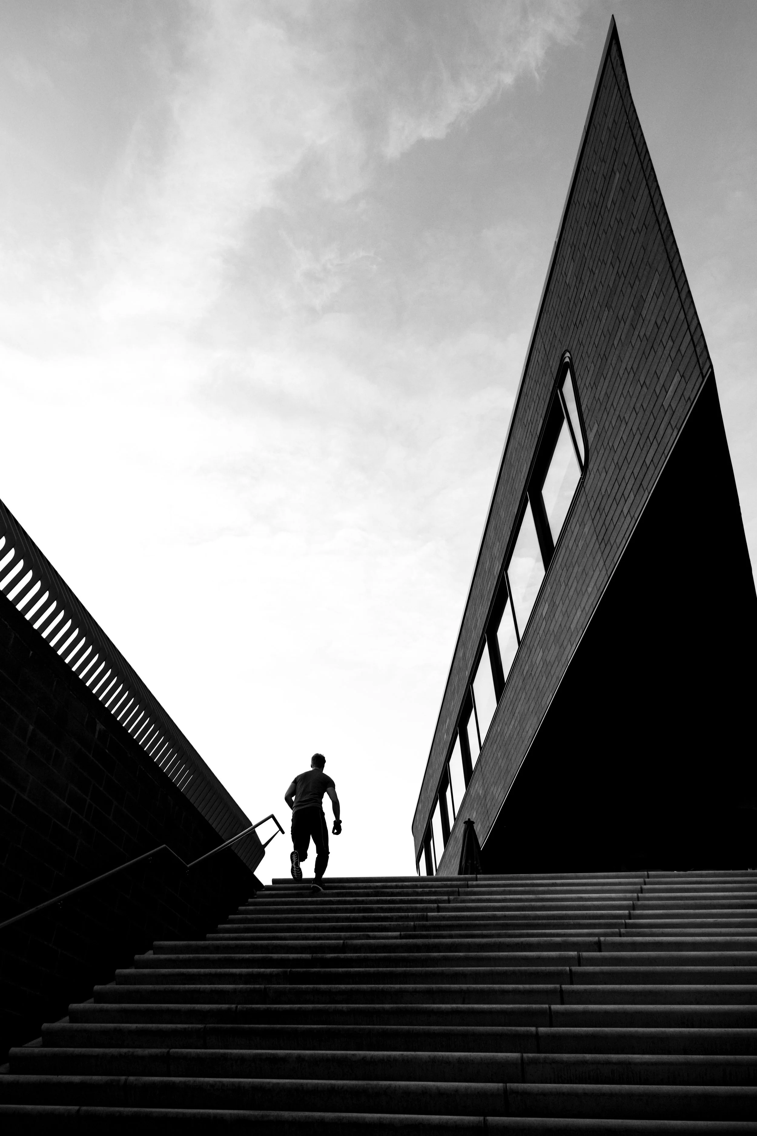 Schwarz-weiß-Fotografie eines Mannes, der die Treppe eines modernen Gebäudes hinaufgeht, mit einem blauen Himmel im Hintergrund.