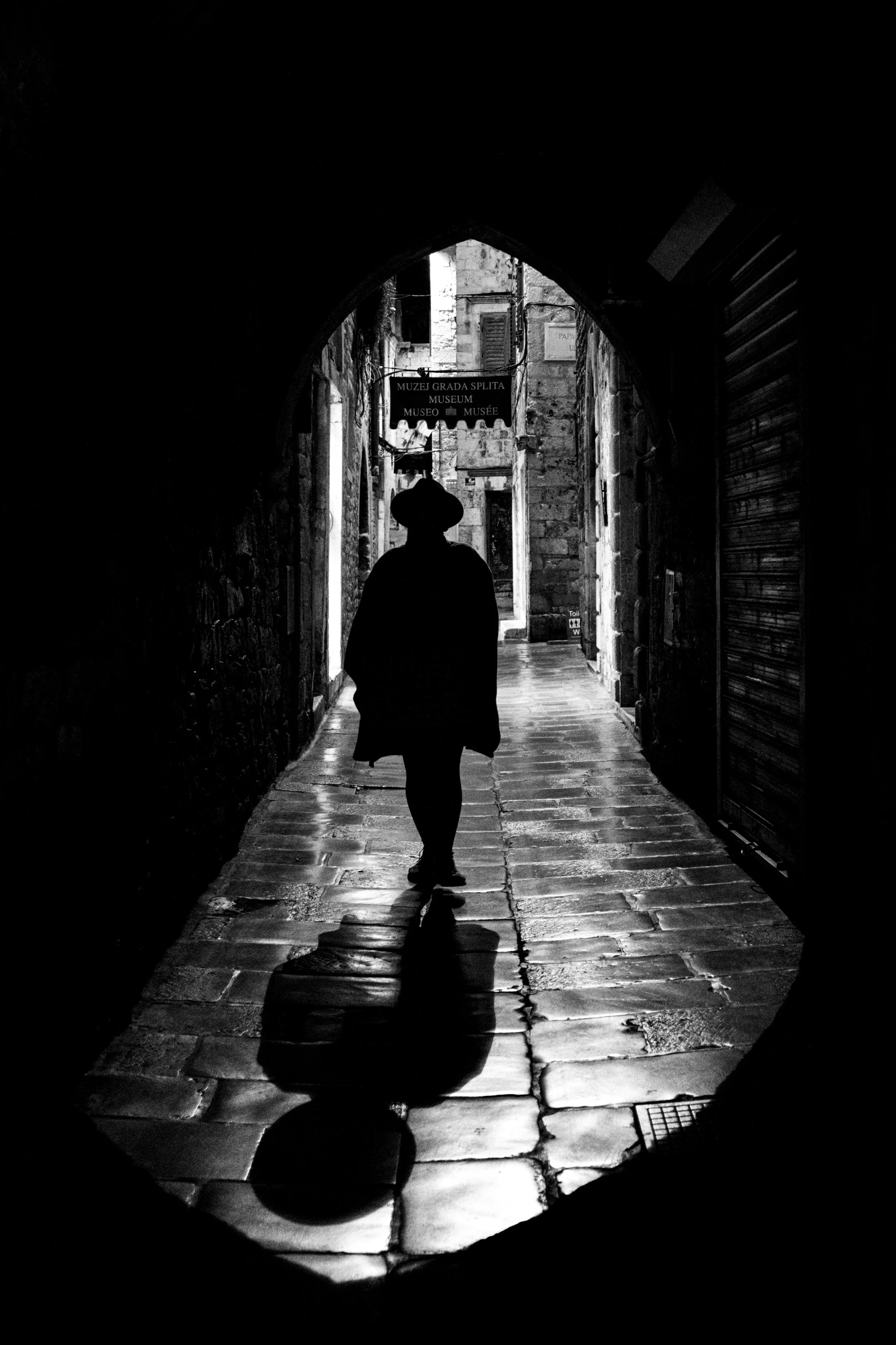 Silhouette einer Person, die durch eine dunkle Gasse in Split, Kroatien, geht, mit nassen Kopfsteinpflaster und einem Schild für das Museum im Hintergrund.