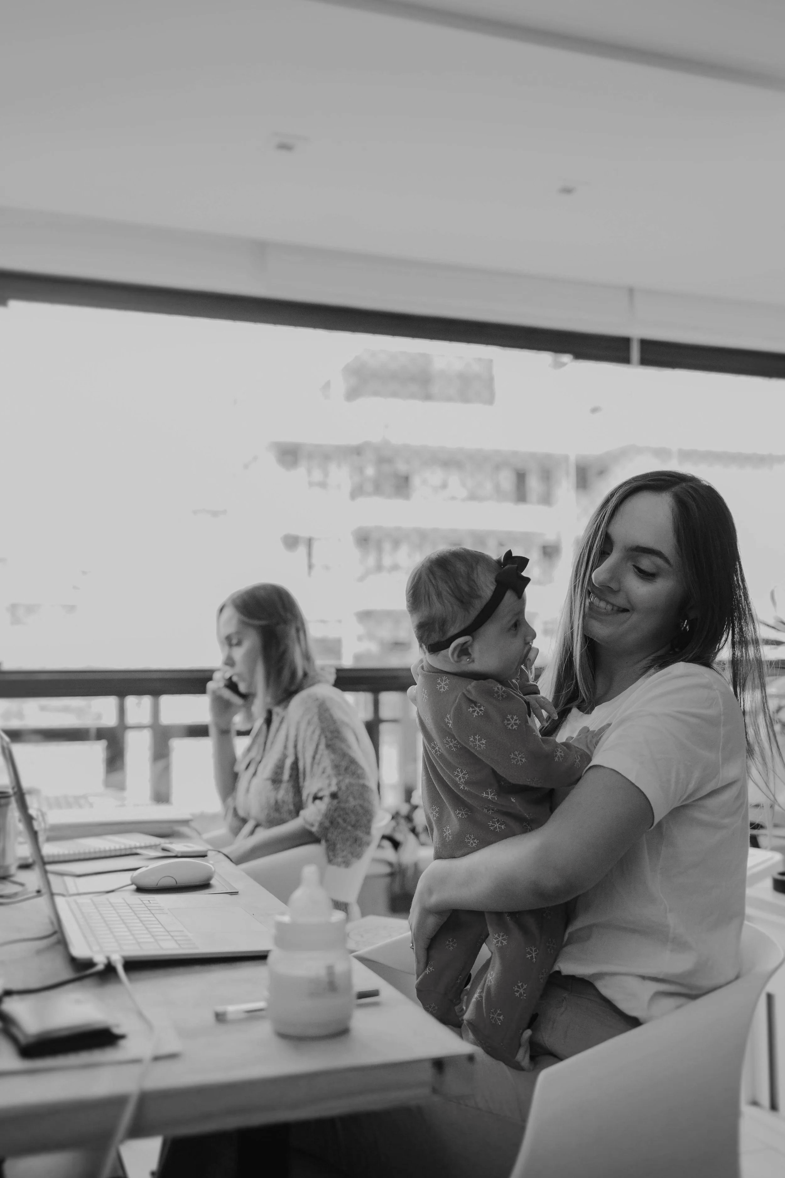 A woman smiling while holding a young girl in her arms in an office environment, with a woman working at a desk in the background.