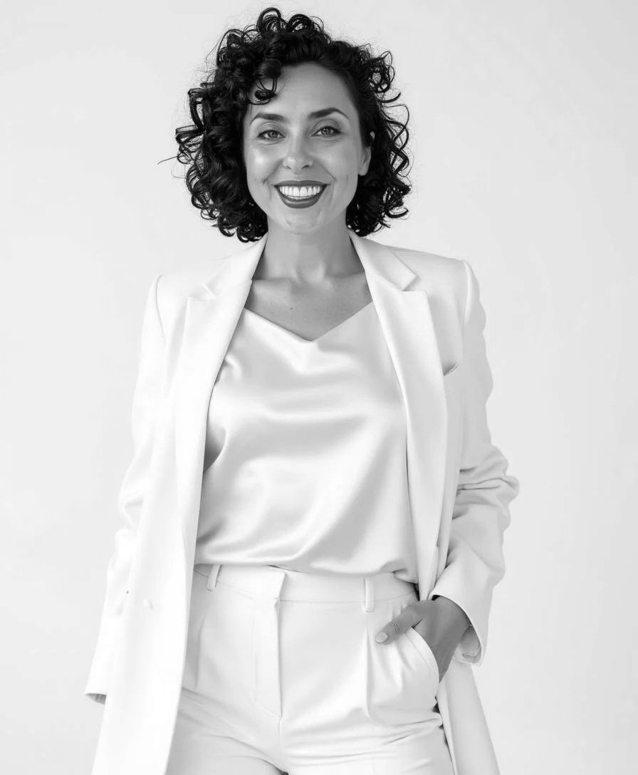 Black and white photo of a woman smiling, wearing a white blazer and silk top, with curly hair, standing against a plain background.