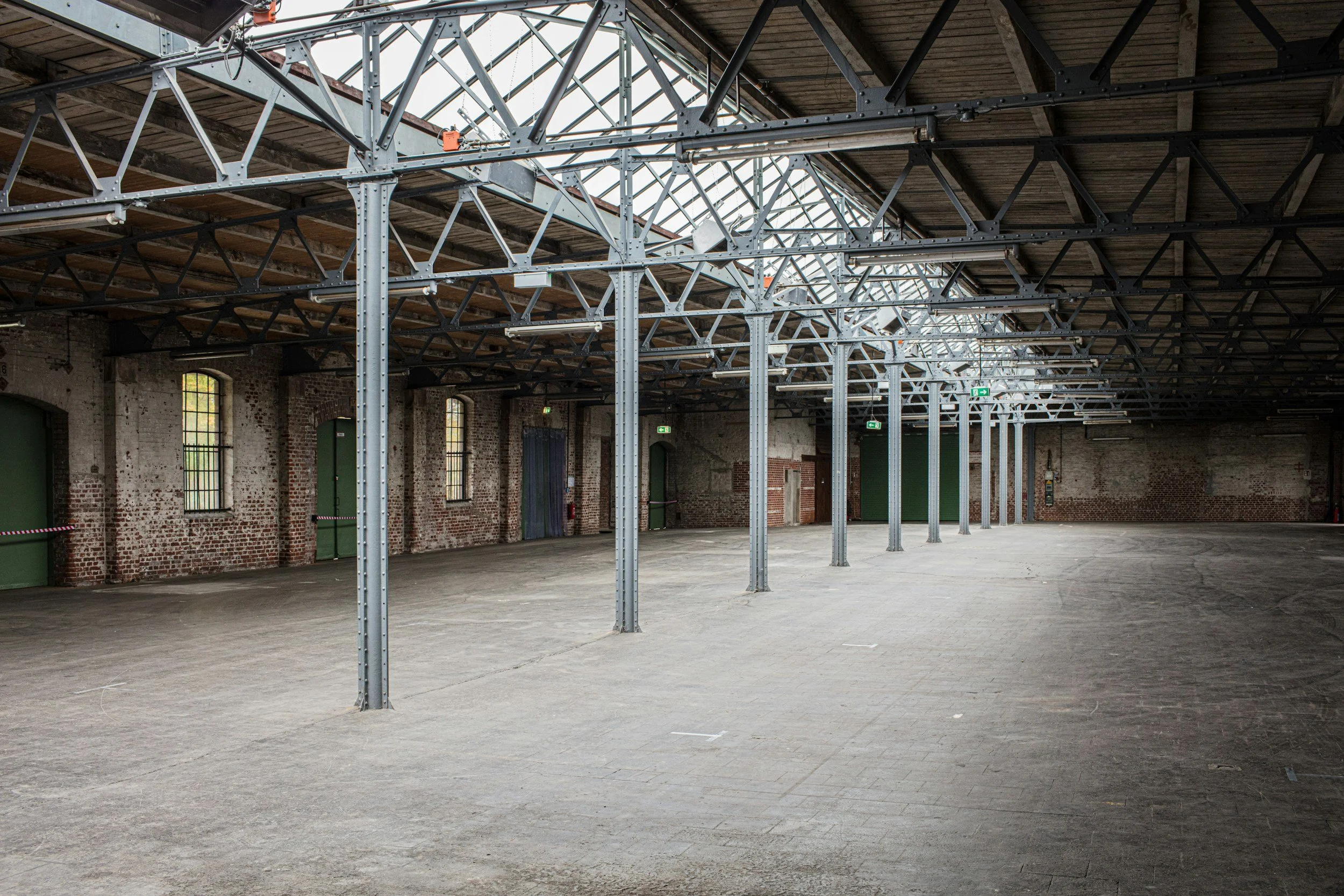 Empty industrial or warehouse space with brick walls, high ceiling, metal support beams, and large windows.
