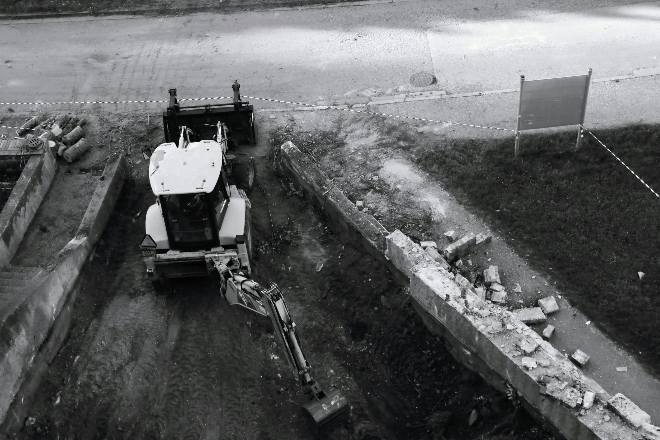 Black and white photo of a construction site with a small excavator working near a partially demolished brick wall, surrounded by construction materials and a street with a sign.