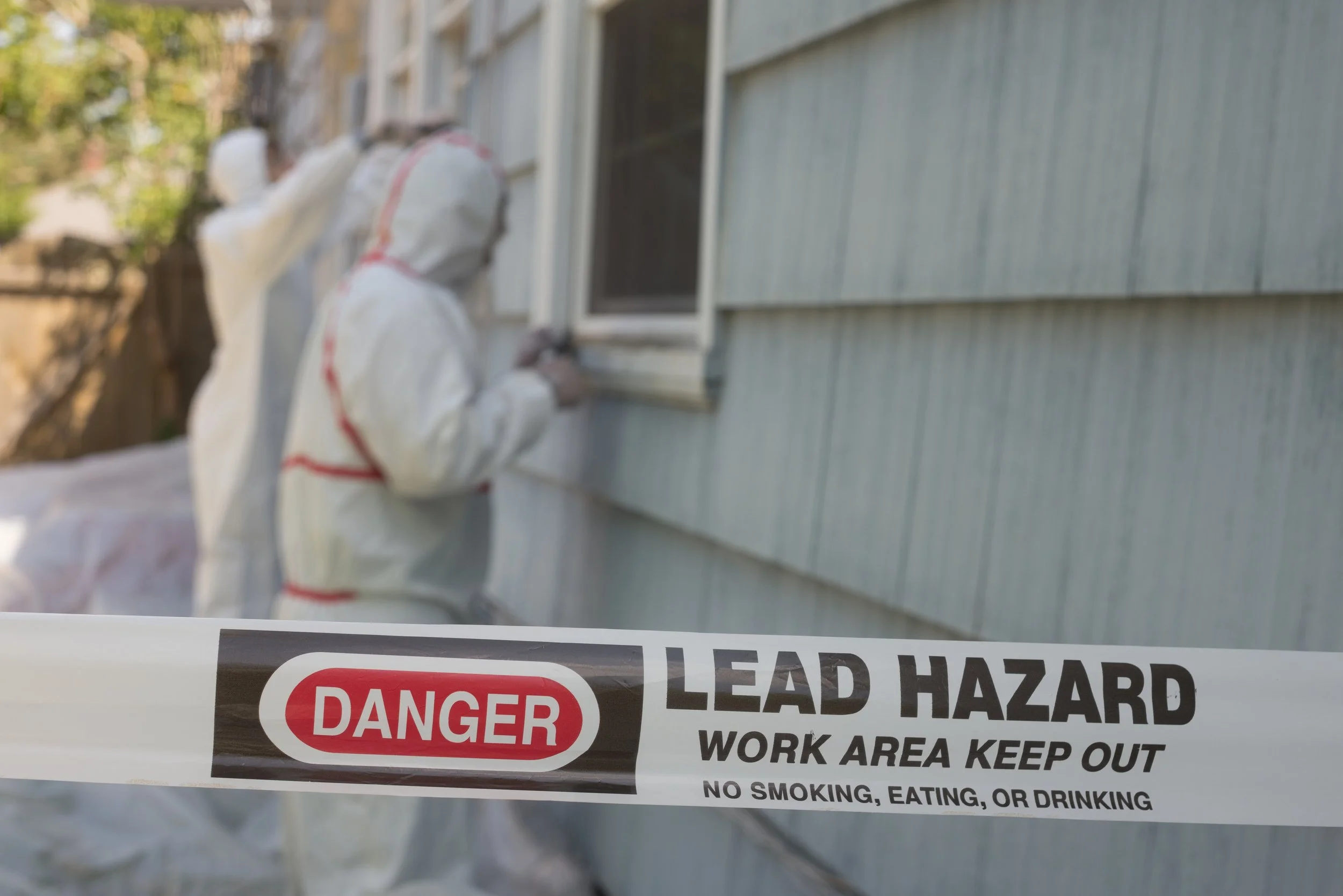 Construction workers in hazmat suits working on the exterior of a house behind caution tape that reads "Danger Lead Hazard Work Area Keep Out" and "No Smoking, Eating, or Drinking."