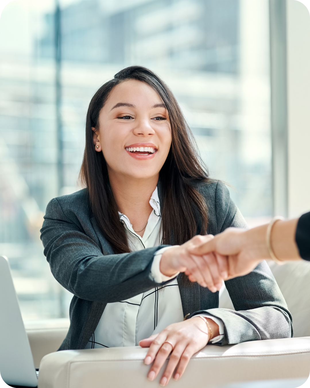 A woman in a business suit smiles as she shakes hands with someone during a business meeting in a modern office.