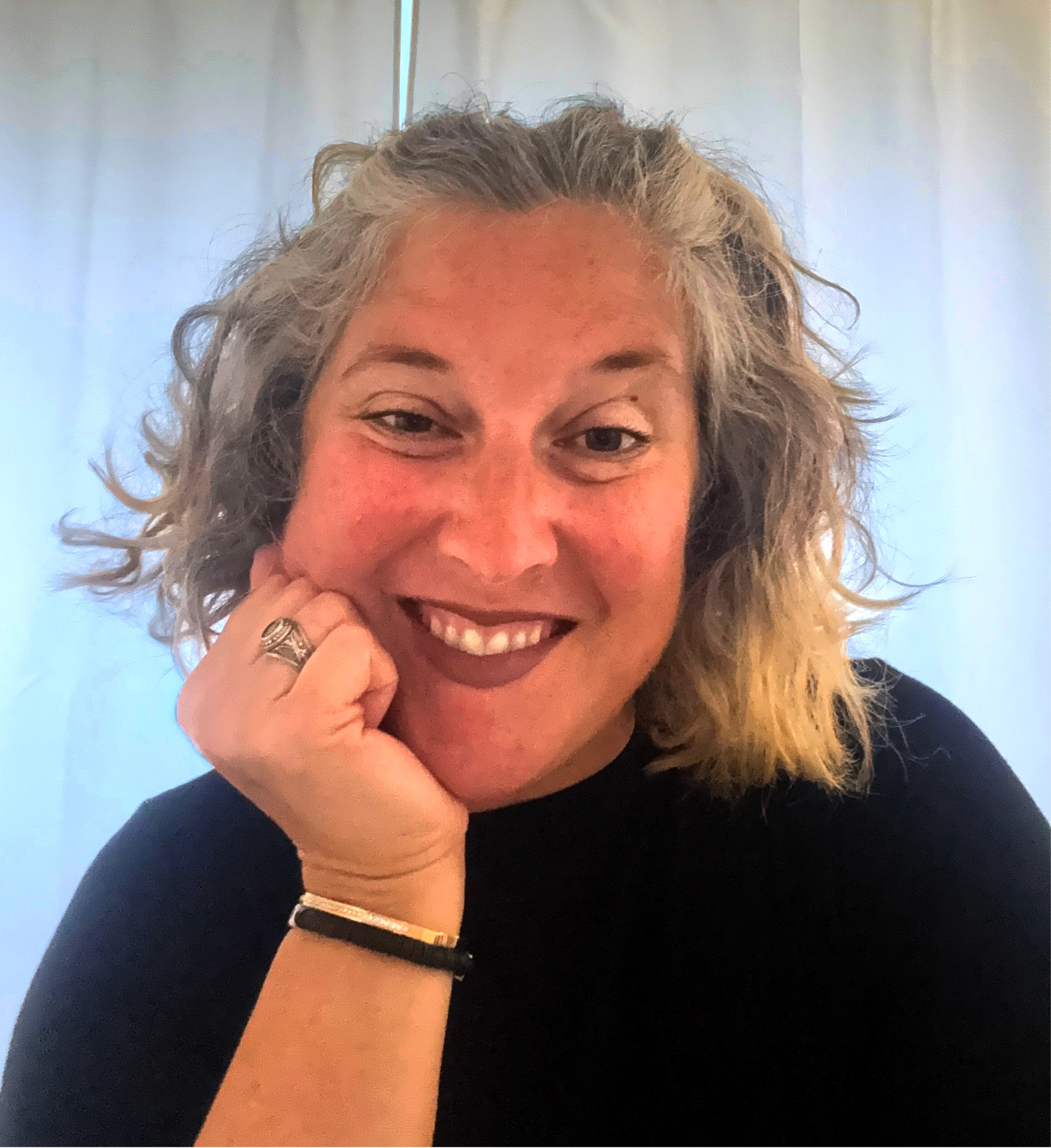 A woman with curly, shoulder-length gray hair smiling with her hand resting on her chin, wearing a black top and a ring on her finger, inside a room with light-colored curtains in the background.