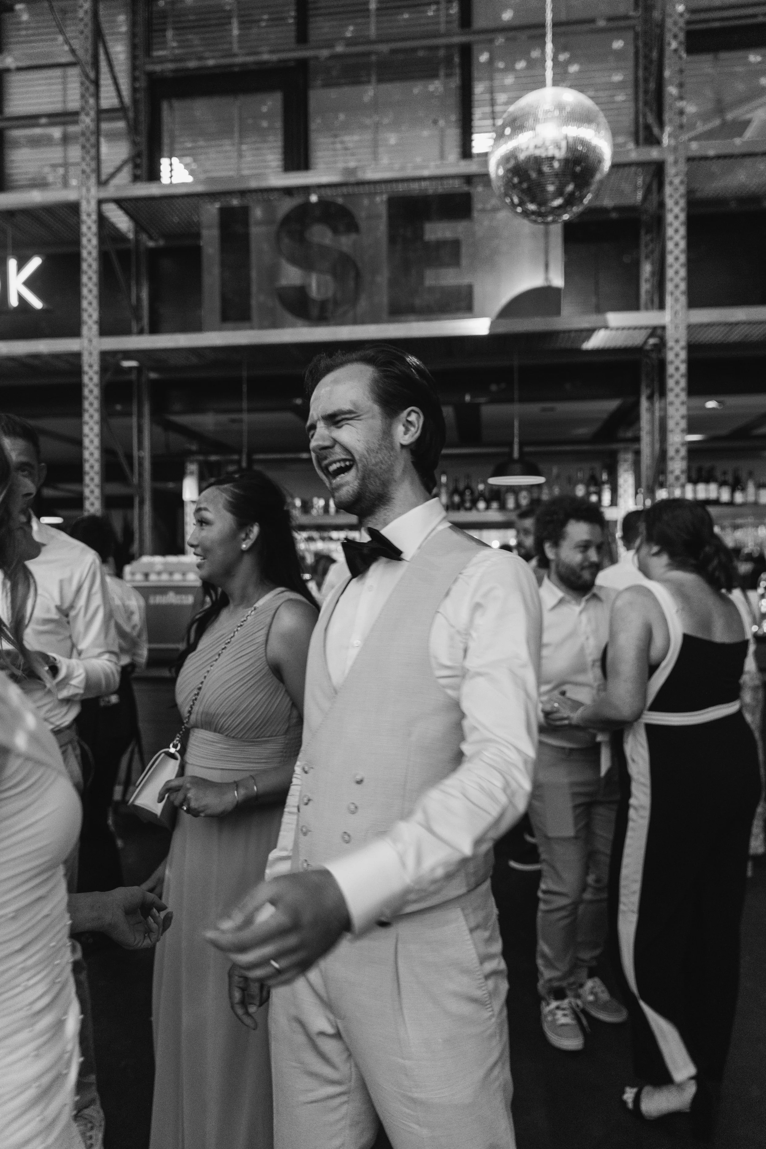 People dressed in formal attire socializing at a social event in a bar or restaurant setting, with a disco ball hanging from the ceiling.