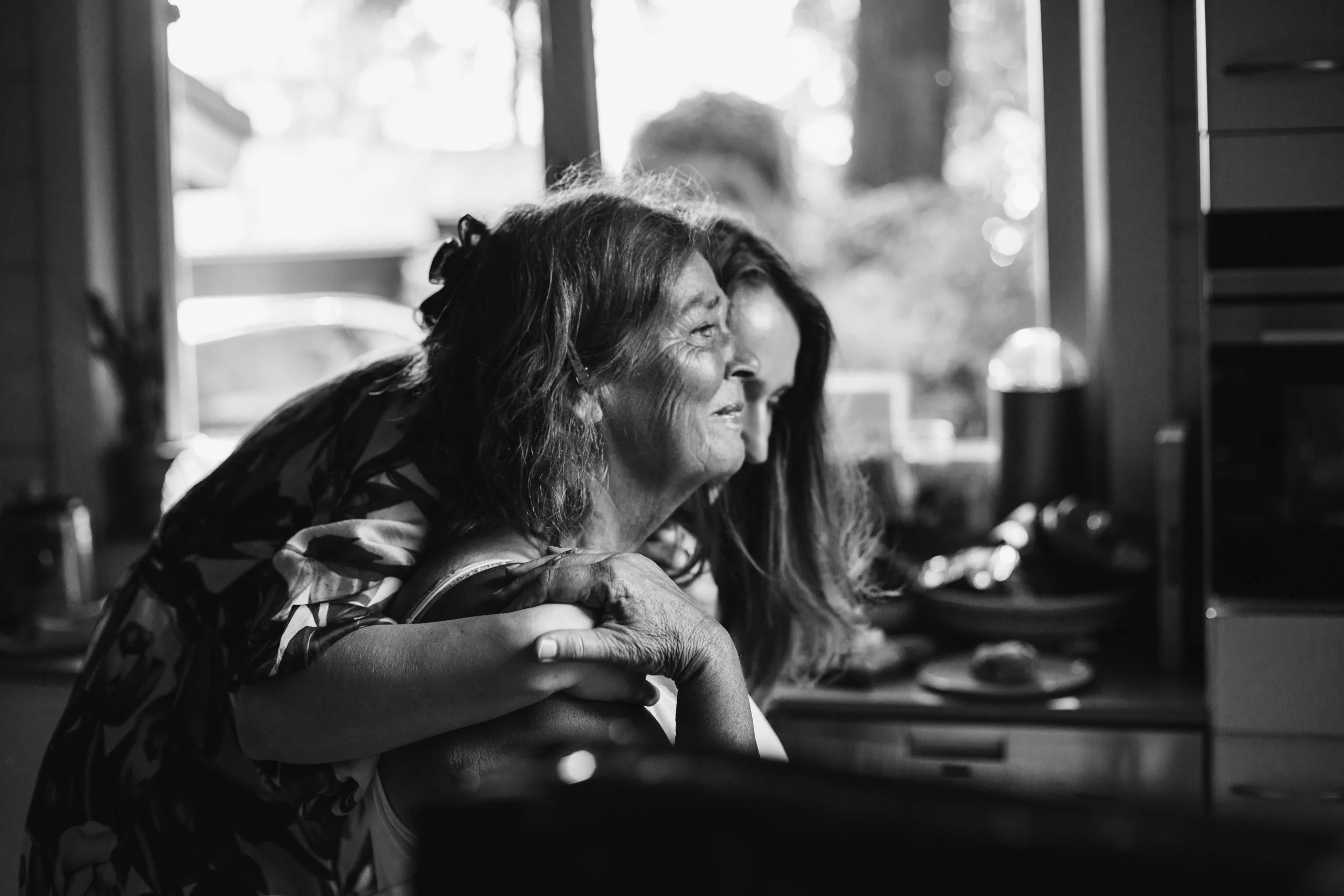 An elderly woman and a younger woman share a moment of affection in a kitchen, with the younger woman hugging the elderly woman from behind. The scene is in black and white.