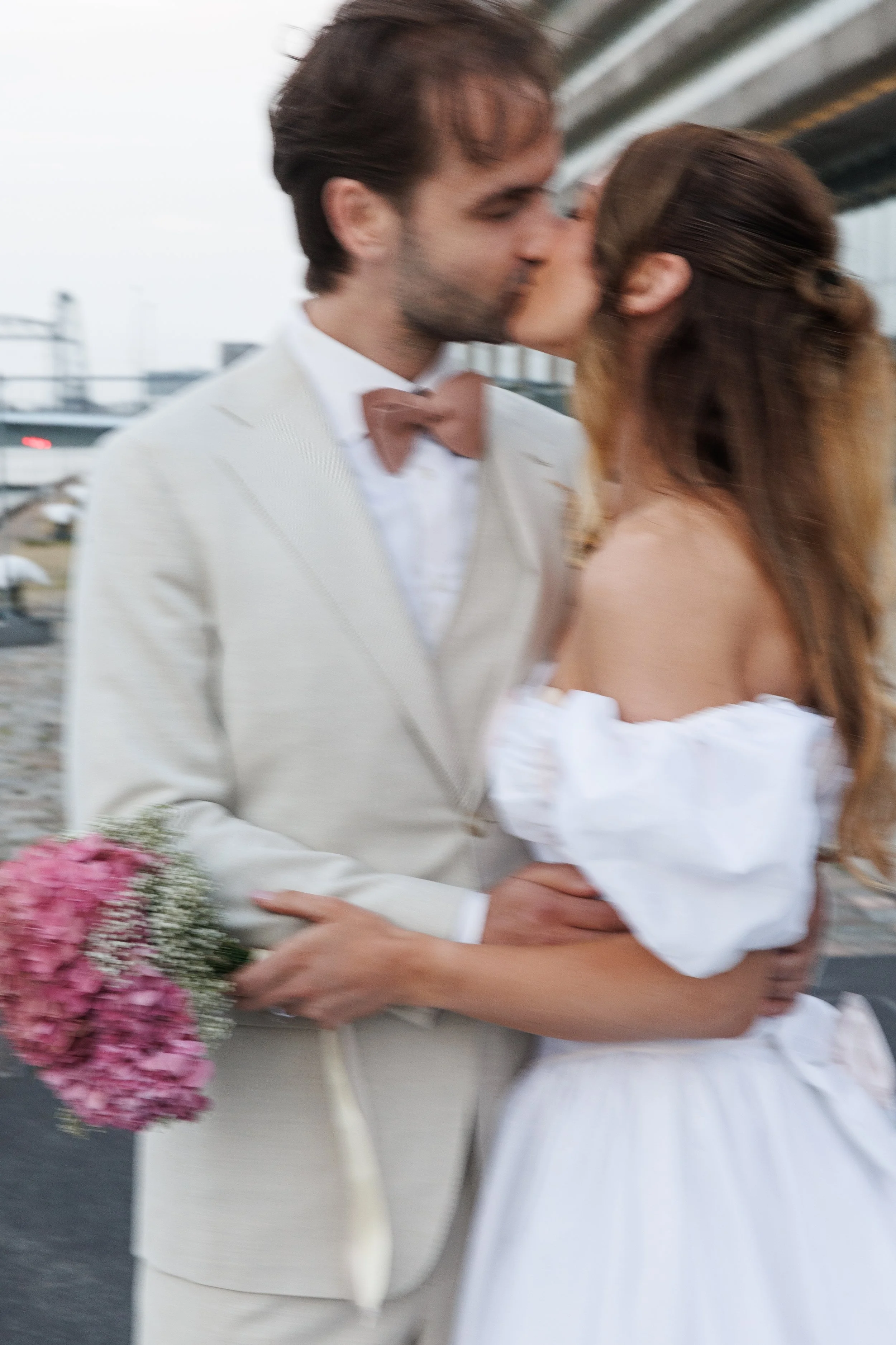 A couple dressed in wedding attire sharing a kiss outdoors, with the man in a white suit and brown bow tie holding a bouquet of pink flowers.