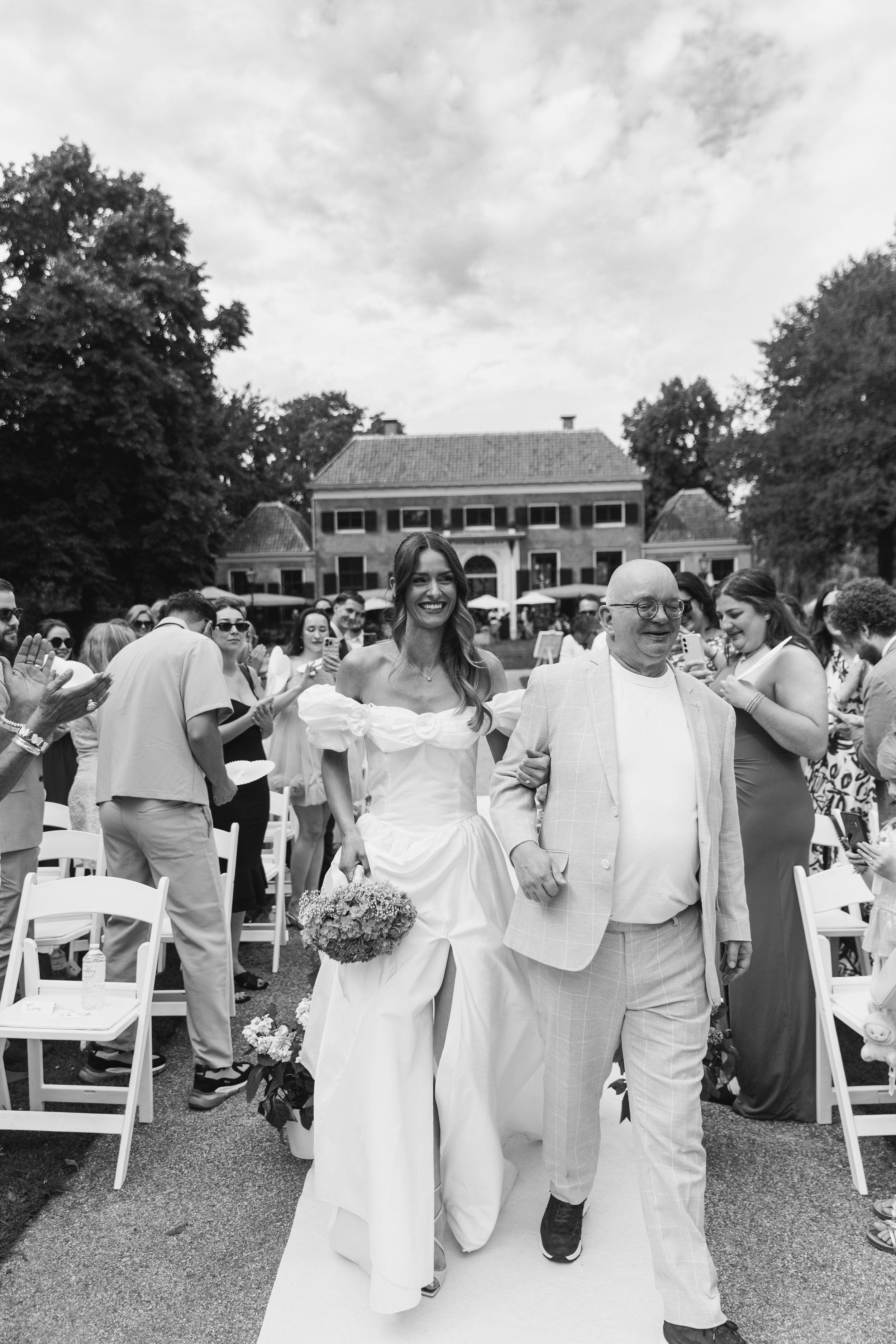 A bride in a white wedding dress walking down the aisle with a man in a checkered suit at an outdoor wedding ceremony, surrounded by guests seated and standing, with large trees and a building in the background.