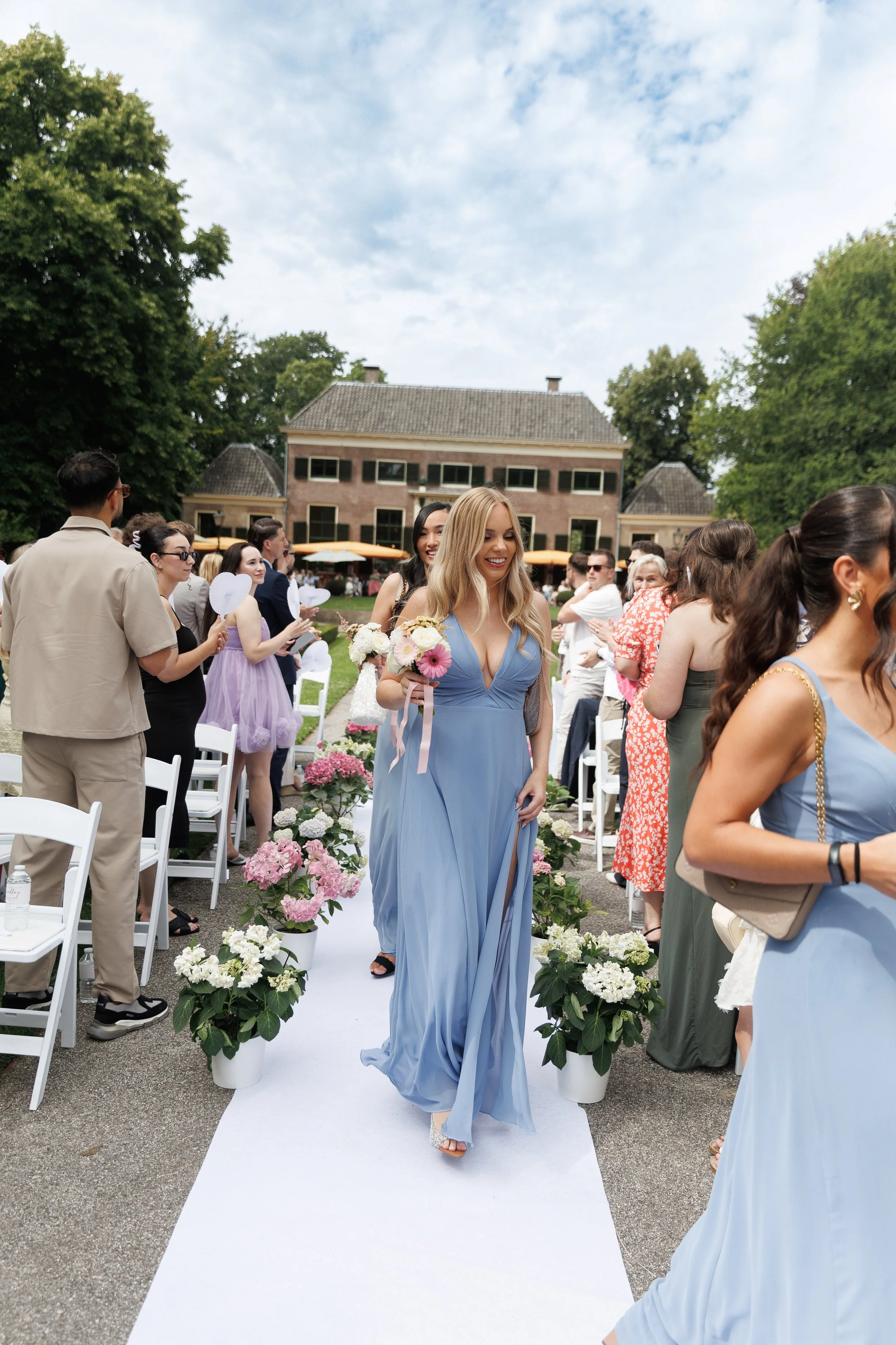 A woman in a blue dress walking down the aisle at an outdoor wedding ceremony, holding a bouquet of flowers, surrounded by guests under a partly cloudy sky.