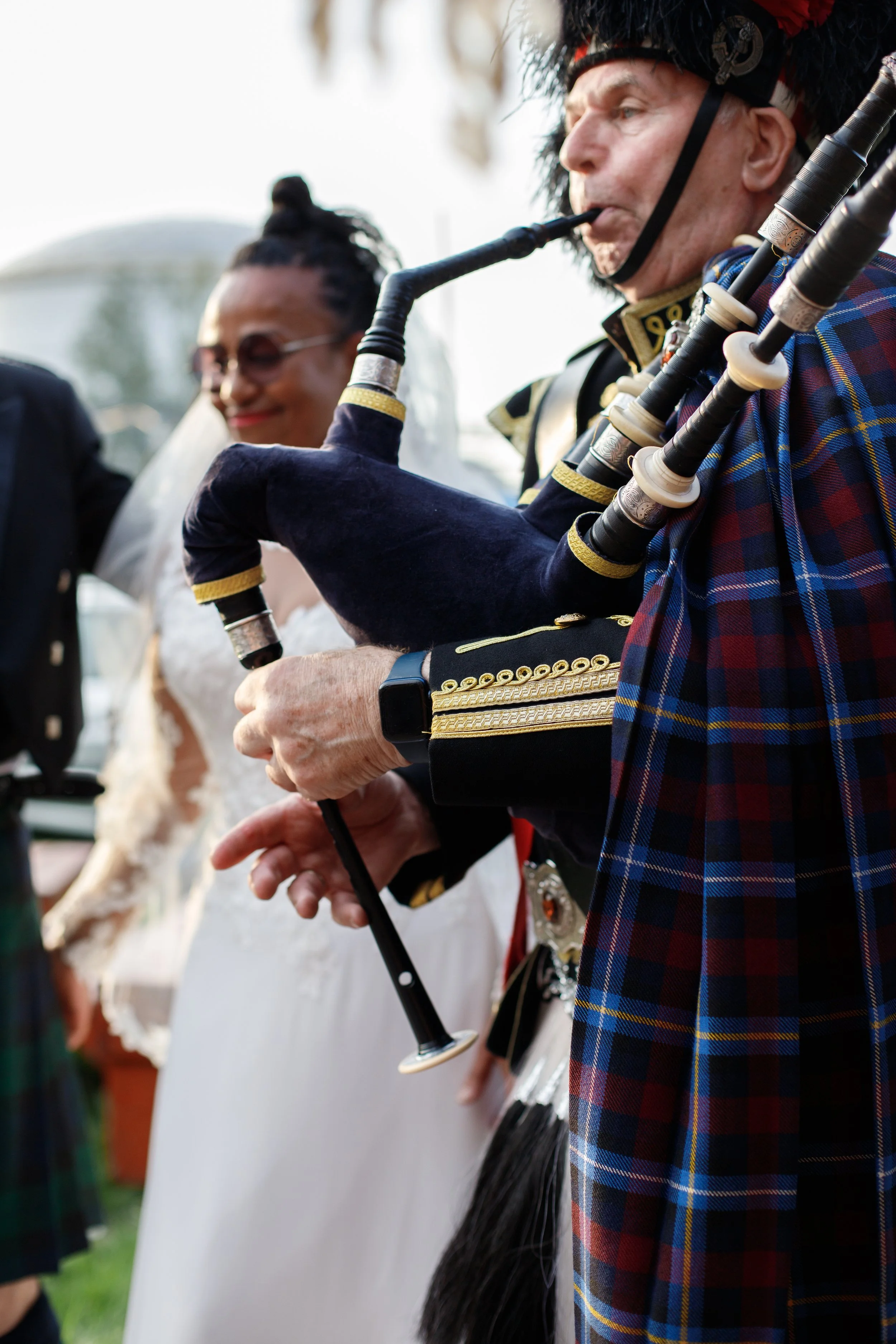 A man in traditional Scottish attire playing bagpipes at an outdoor event, with a woman in a wedding dress smiling in the background.