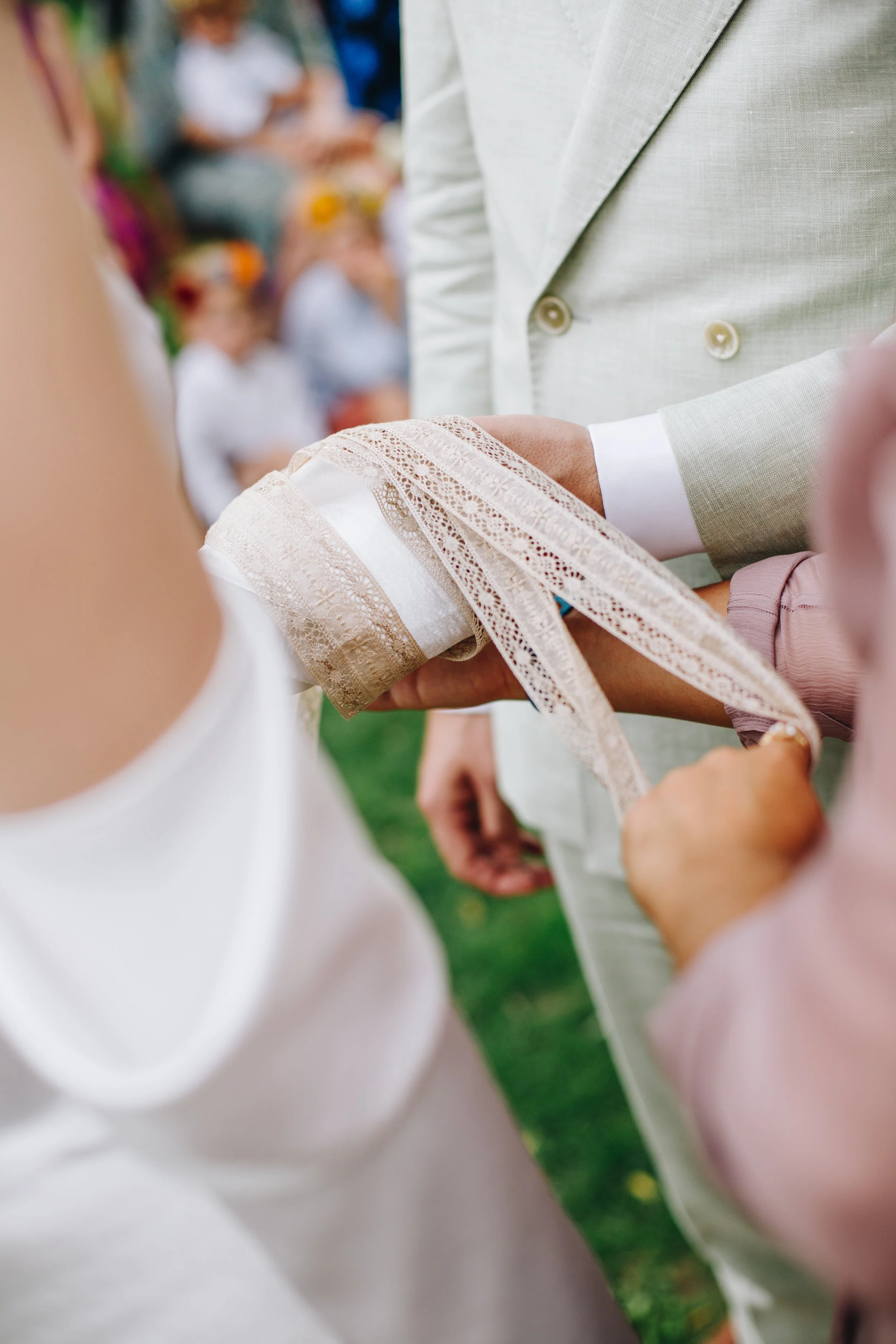 Close-up of a wedding ceremony with a groom tying a lace ribbon around the bride's wrist, with guests blurred in the background.