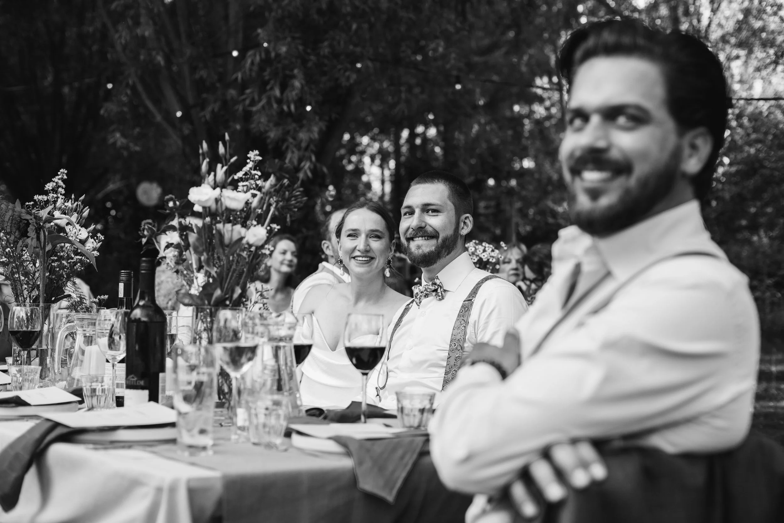 A group of people sitting at a table outdoors during a celebration or wedding, smiling and enjoying drinks, with flowers and wine bottles on the table, in black and white.