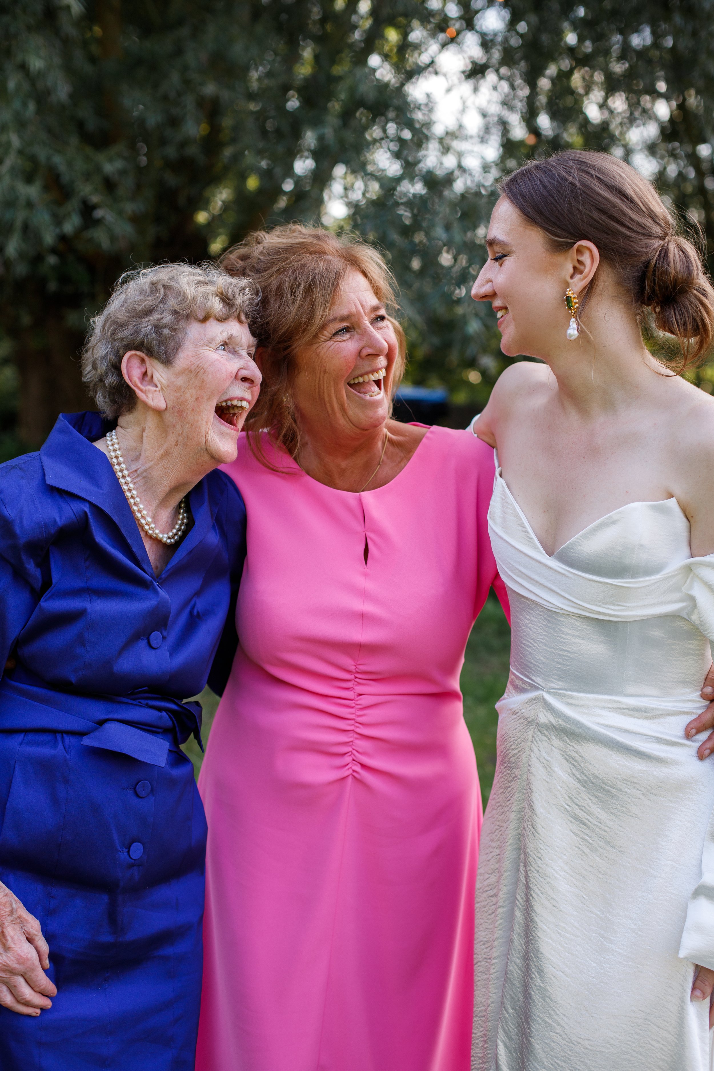 Three women, an elderly woman in a blue dress, a middle-aged woman in a pink dress, and a young woman in a white dress, are outdoors laughing and smiling together.