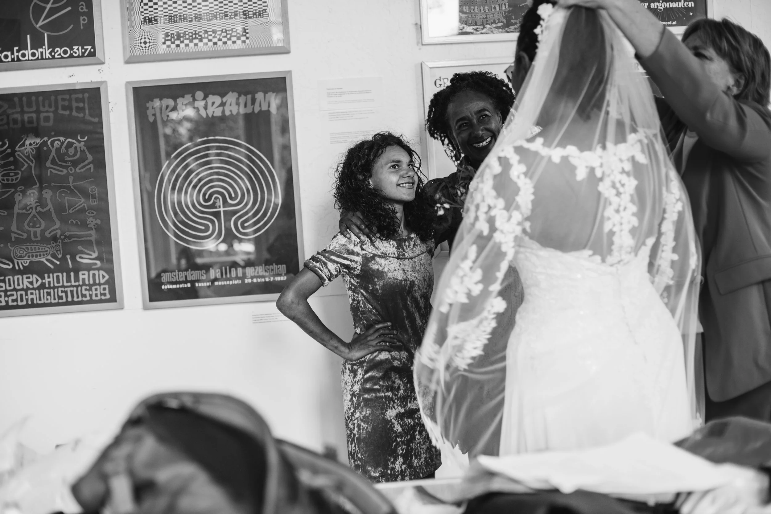 A bride in a wedding dress with a veil is surrounded by three women, smiling and talking. One woman has curly hair and is wearing a patterned dress. The scene is indoors, with framed posters on the wall in the background.