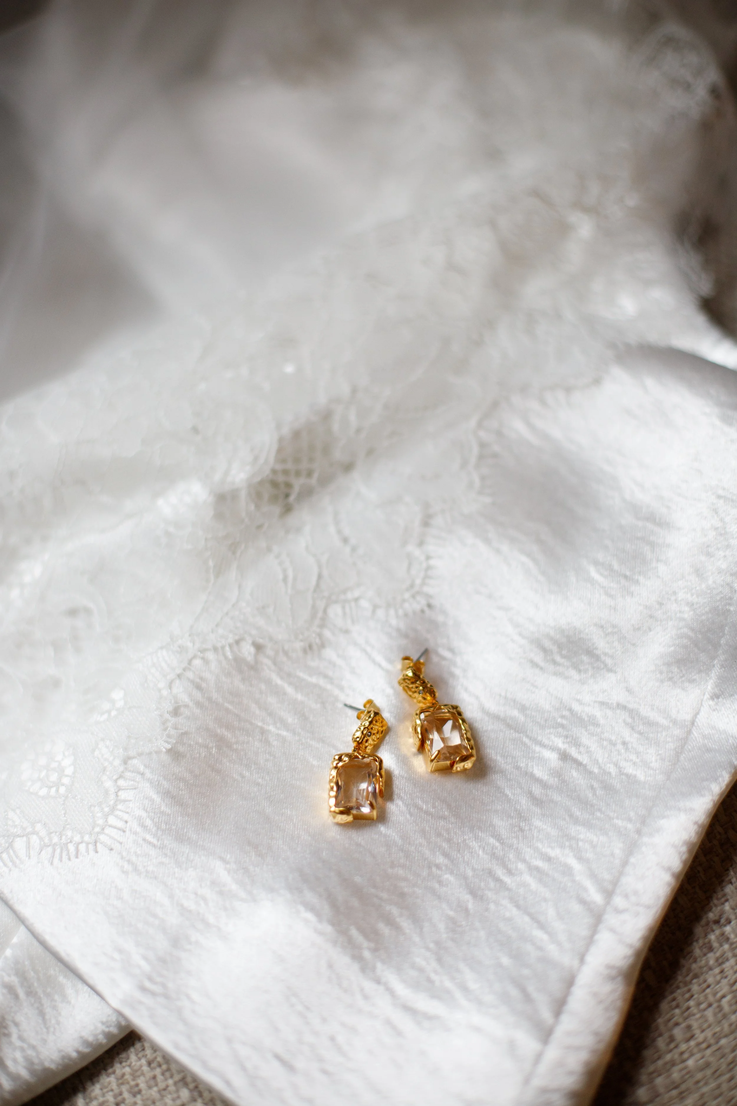 Gold earrings with large rectangular clear gemstones resting on white satin fabric, possibly part of bridal jewelry.