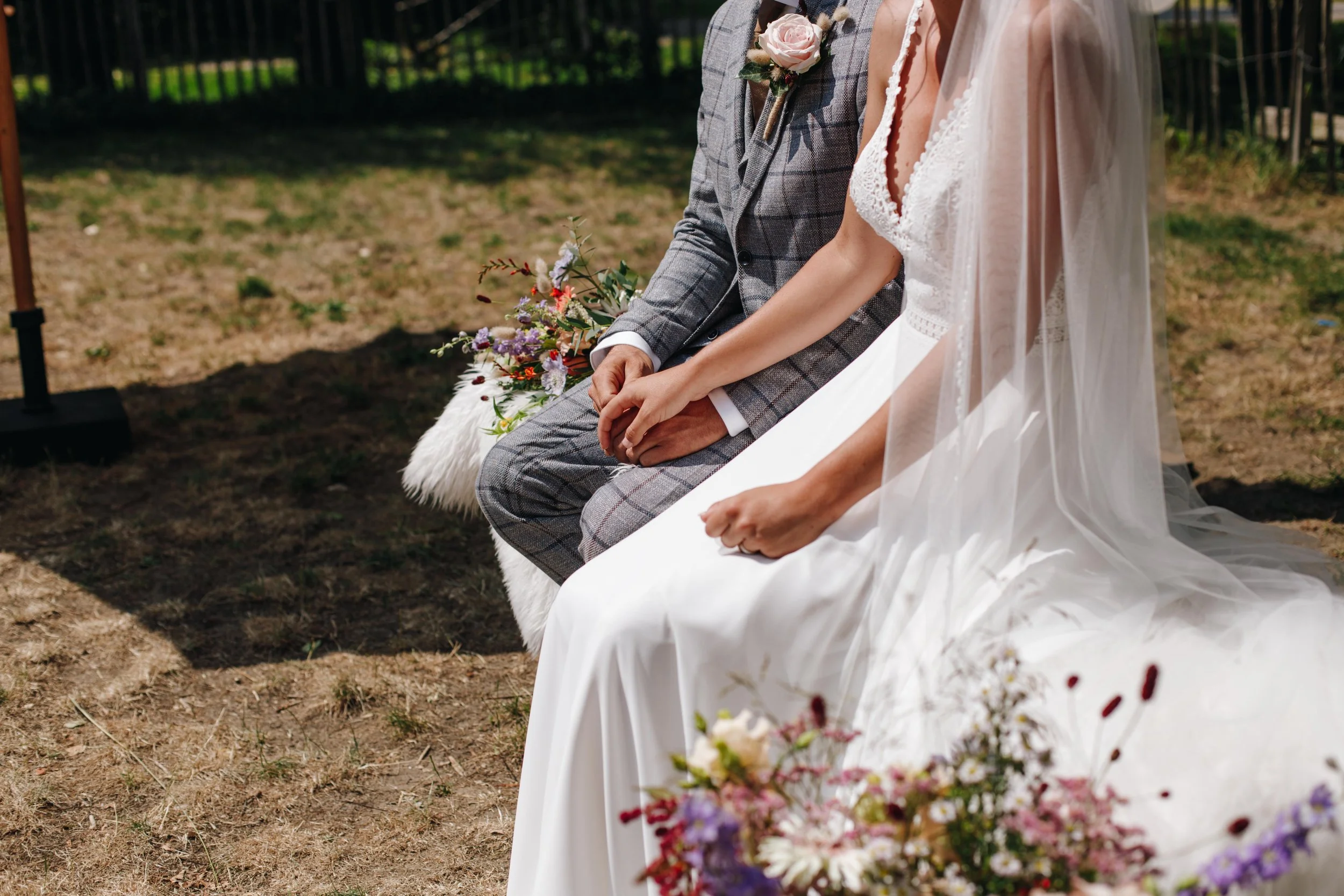 A bride and groom sitting outdoors on a dirt surface during their wedding ceremony, holding hands. The bride is dressed in a white wedding gown with a veil, and the groom is wearing a gray plaid suit with a pink boutonnière. There are colorful flower
