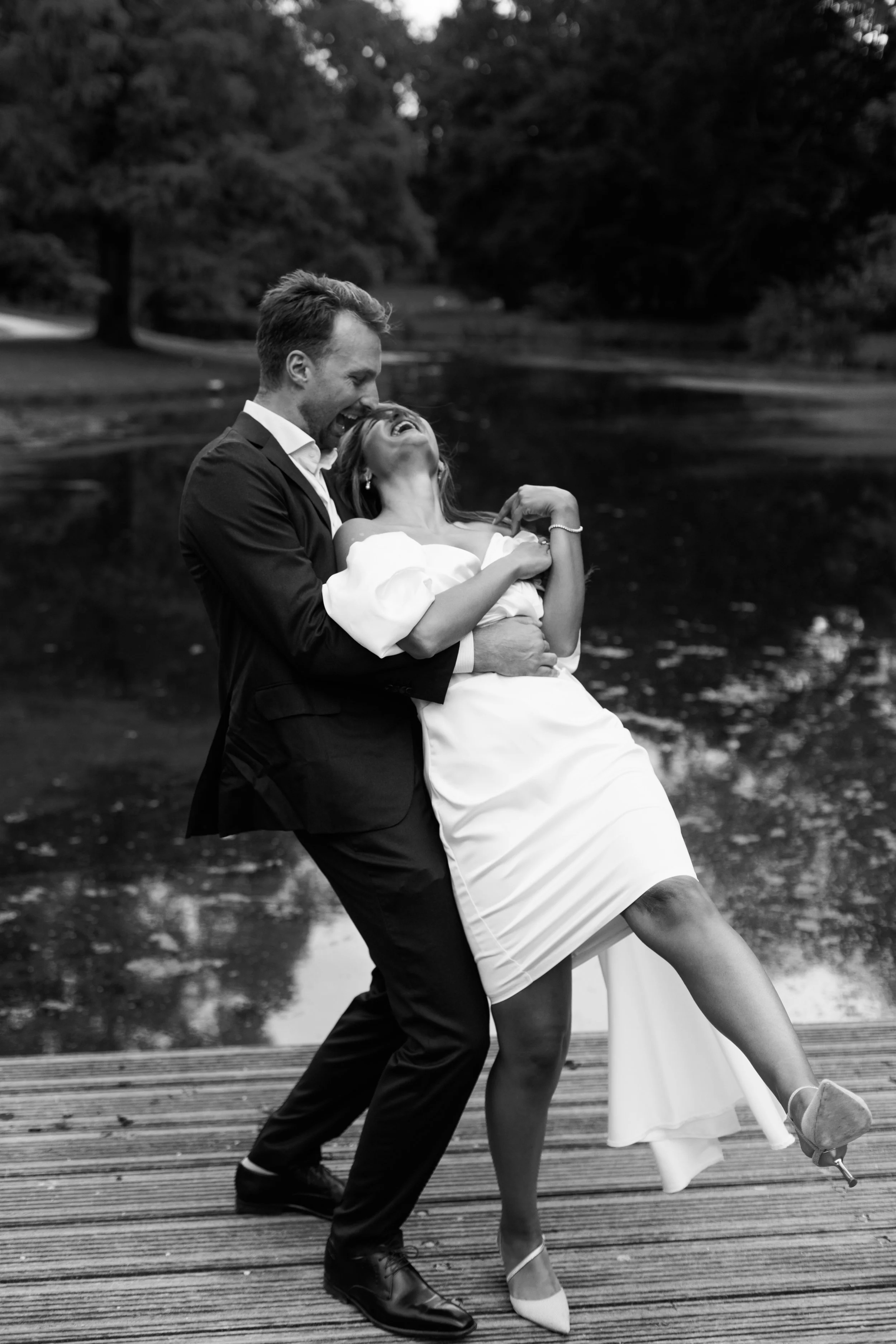 A black and white photo of a joyful couple embracing and laughing on a wooden dock near a pond or lake in a park.