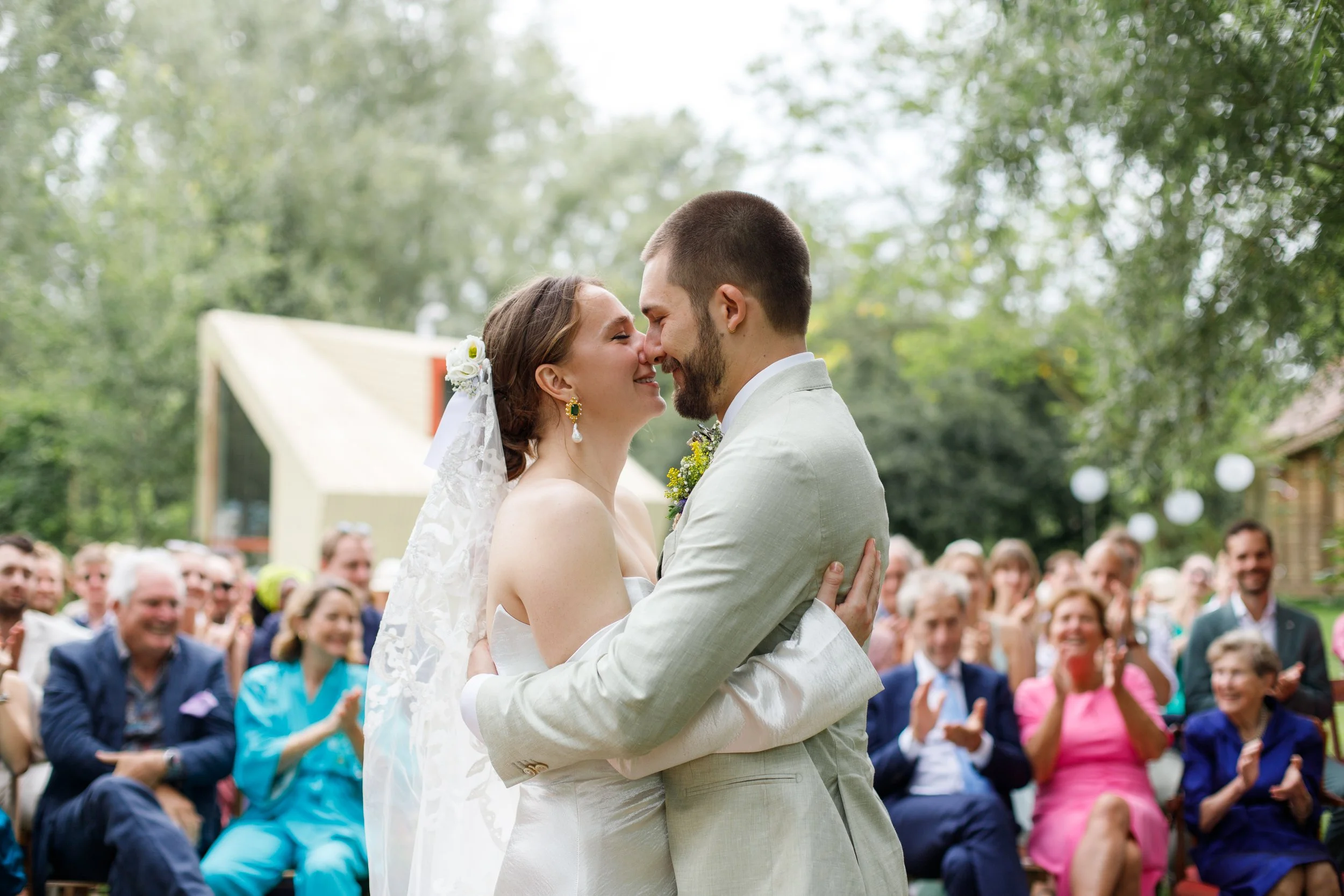 Bride and groom hugging at their wedding ceremony outdoors, with guests clapping and smiling in the background.