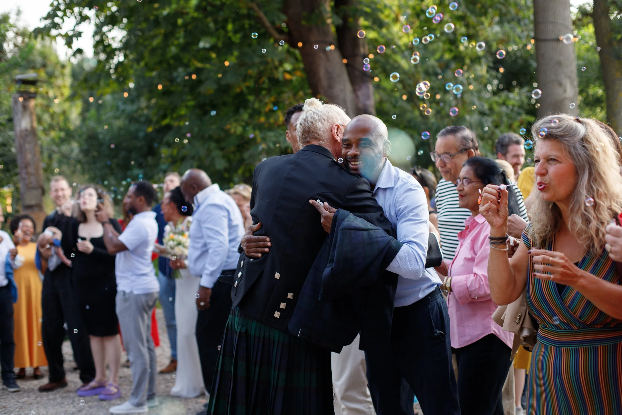 People celebrating outdoors, hugging, blowing bubbles, and smiling in a park or garden.