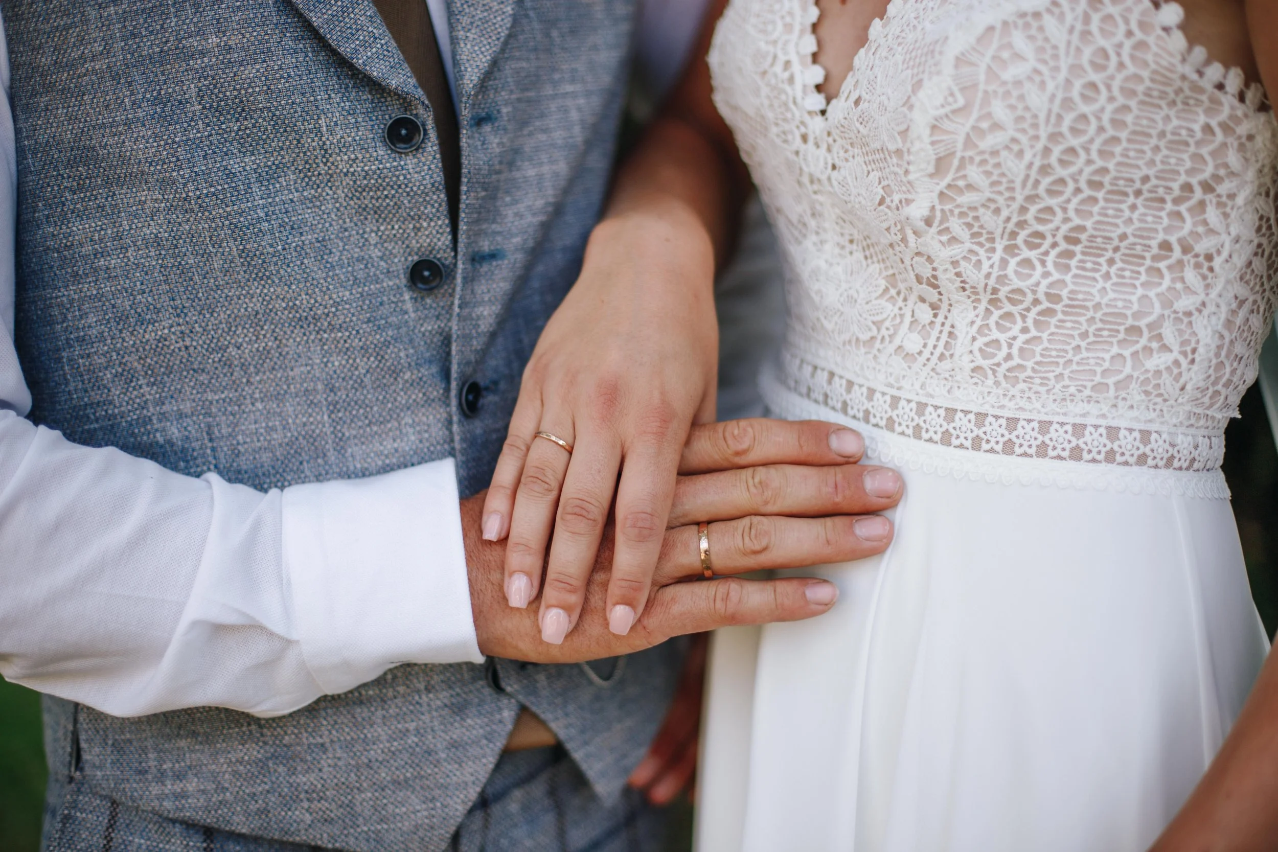 Close-up of a bride and groom holding hands, showing wedding rings, with the groom in a gray vest and white shirt, and the bride in a white lace dress.