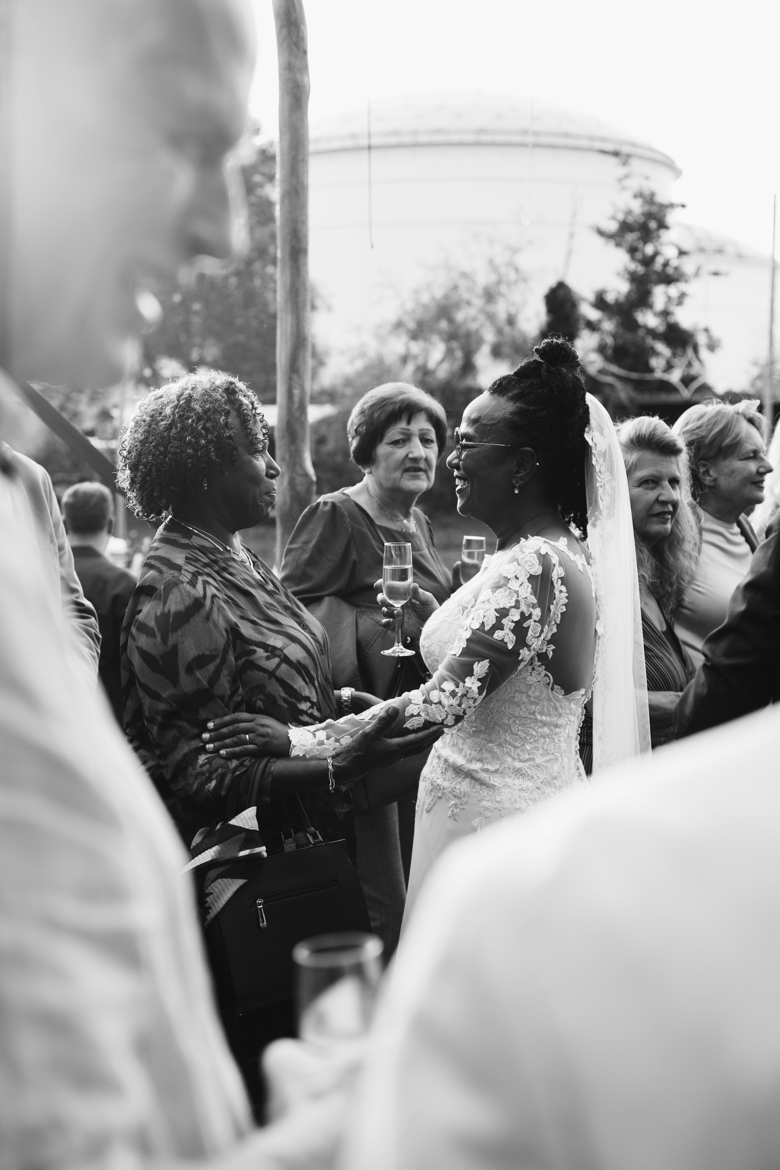 A bride in a lace wedding dress and veil is conversing with an older woman in a patterned blouse at an outdoor wedding reception, with other guests holding glasses of champagne in the background.