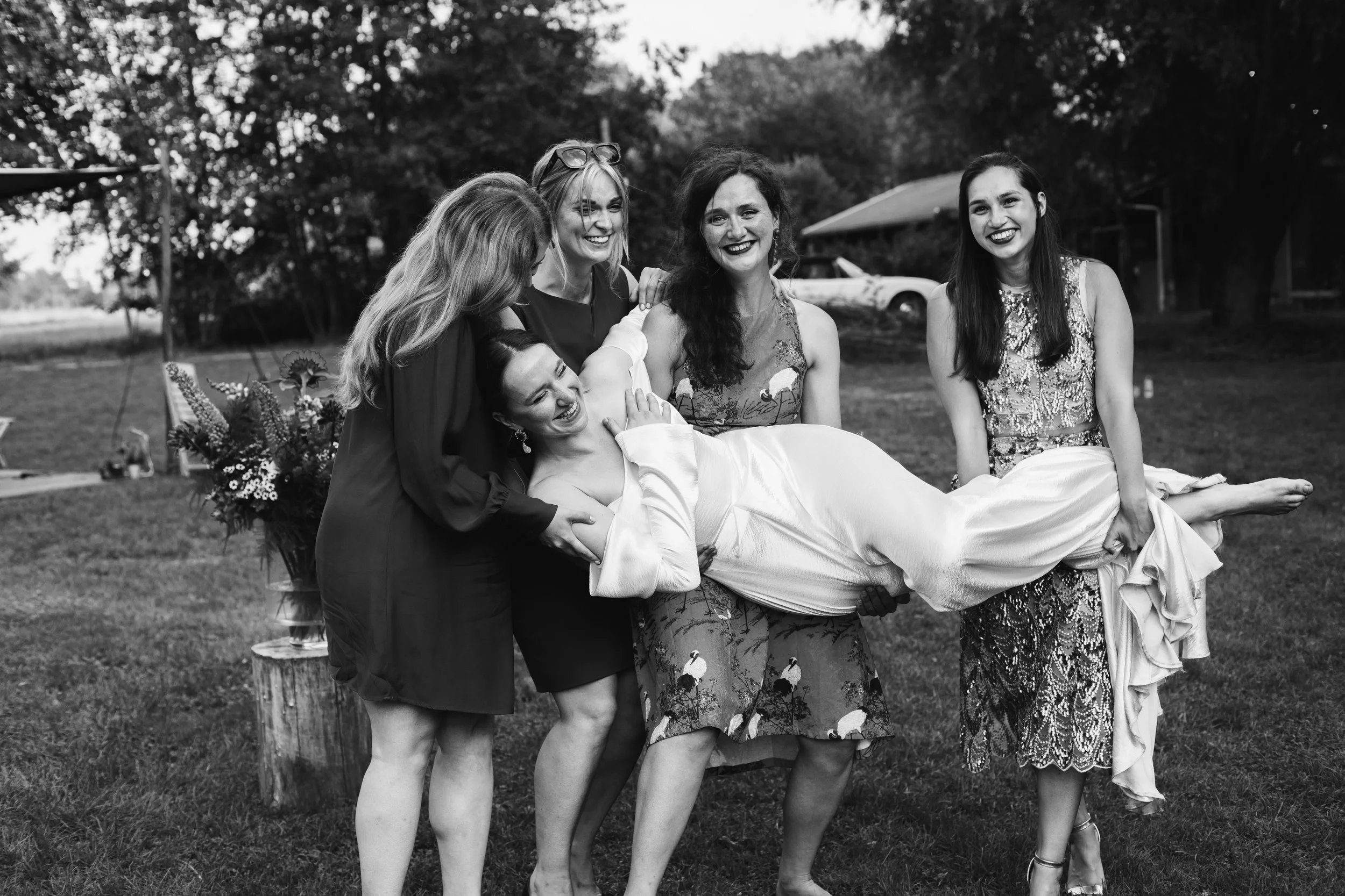 A group of women are playfully holding a woman dressed in a wedding gown, smiling and laughing outdoors in a grassy area with trees and a small building in the background.