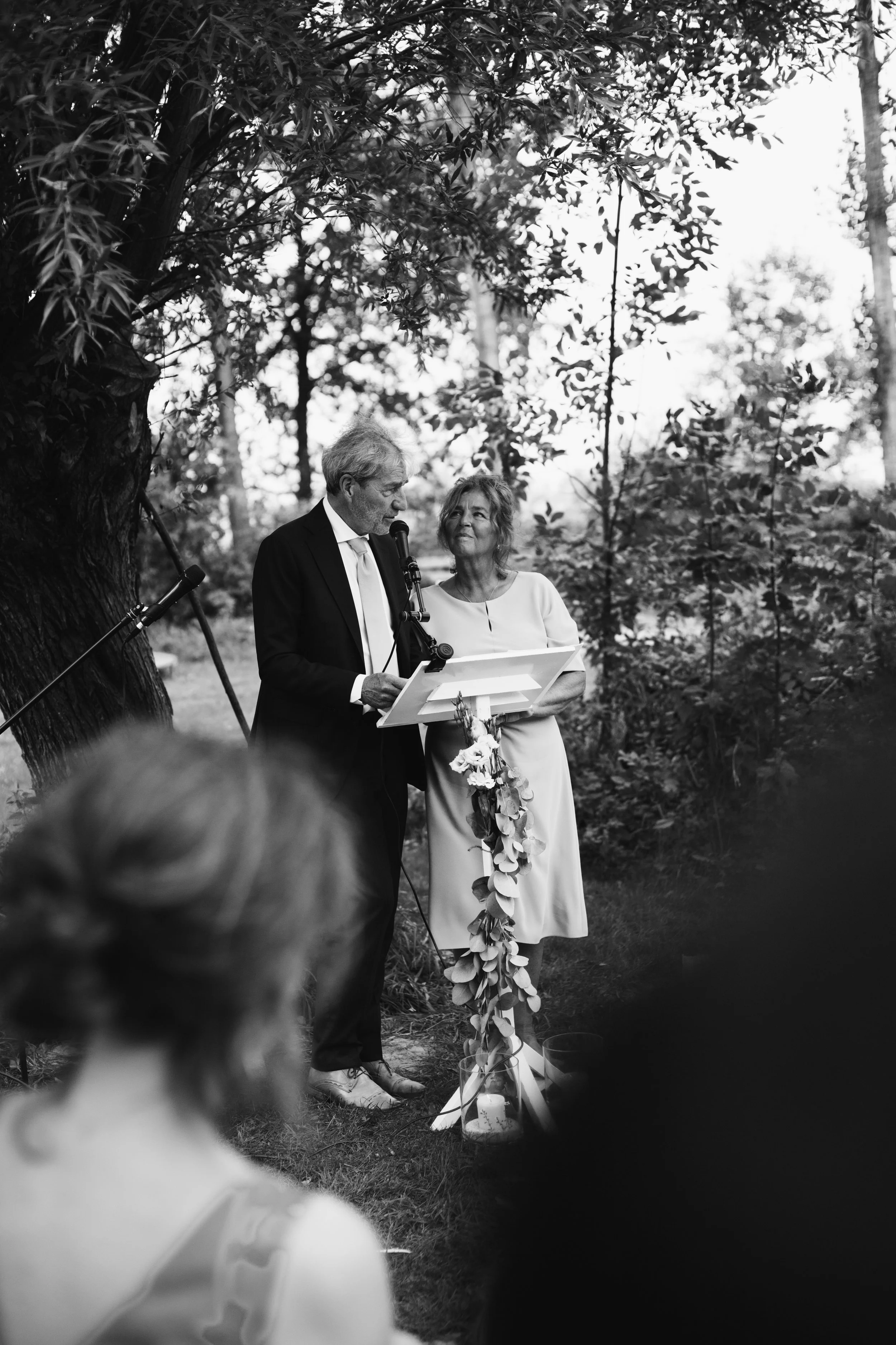A black and white photograph of a wedding ceremony taking place outdoors under a large tree. A man in a suit and a woman in a dress are standing together in front of a lectern, reading vows or giving a speech. The scene is surrounded by natural folia