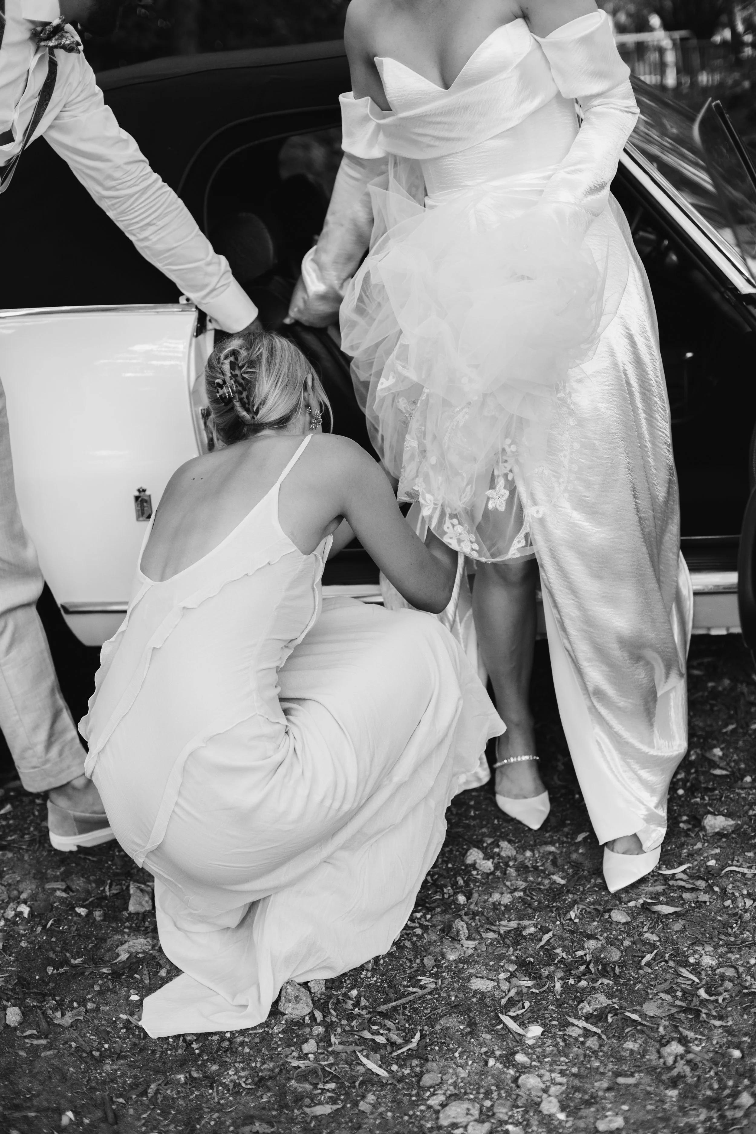 A woman in a wedding dress is helping another woman, dressed in white, into a car. A man is nearby, assisting with the process. The scene appears to be part of a wedding celebration.