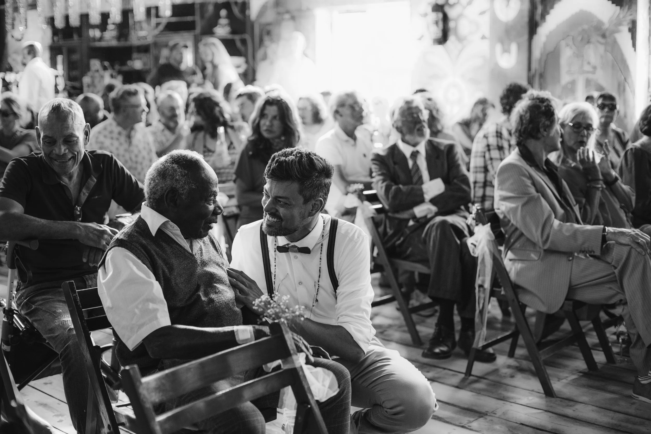 People attending a social gathering or event, seated indoors, with some engaged in conversation and others looking around, in black and white.