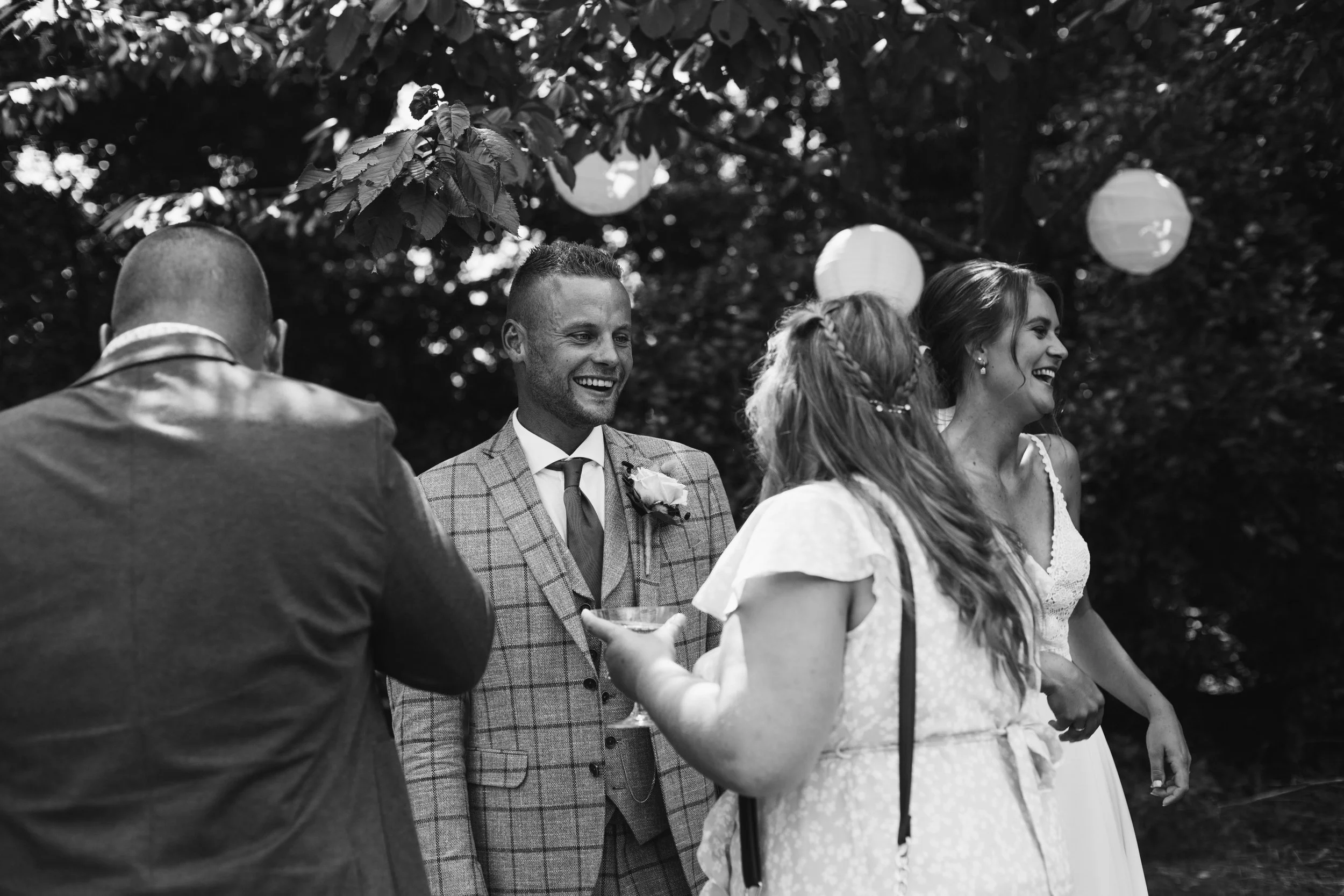 Black and white photo of a wedding celebration outdoors. A bride and groom are smiling, surrounded by guests, with lanterns hanging overhead and trees in the background.