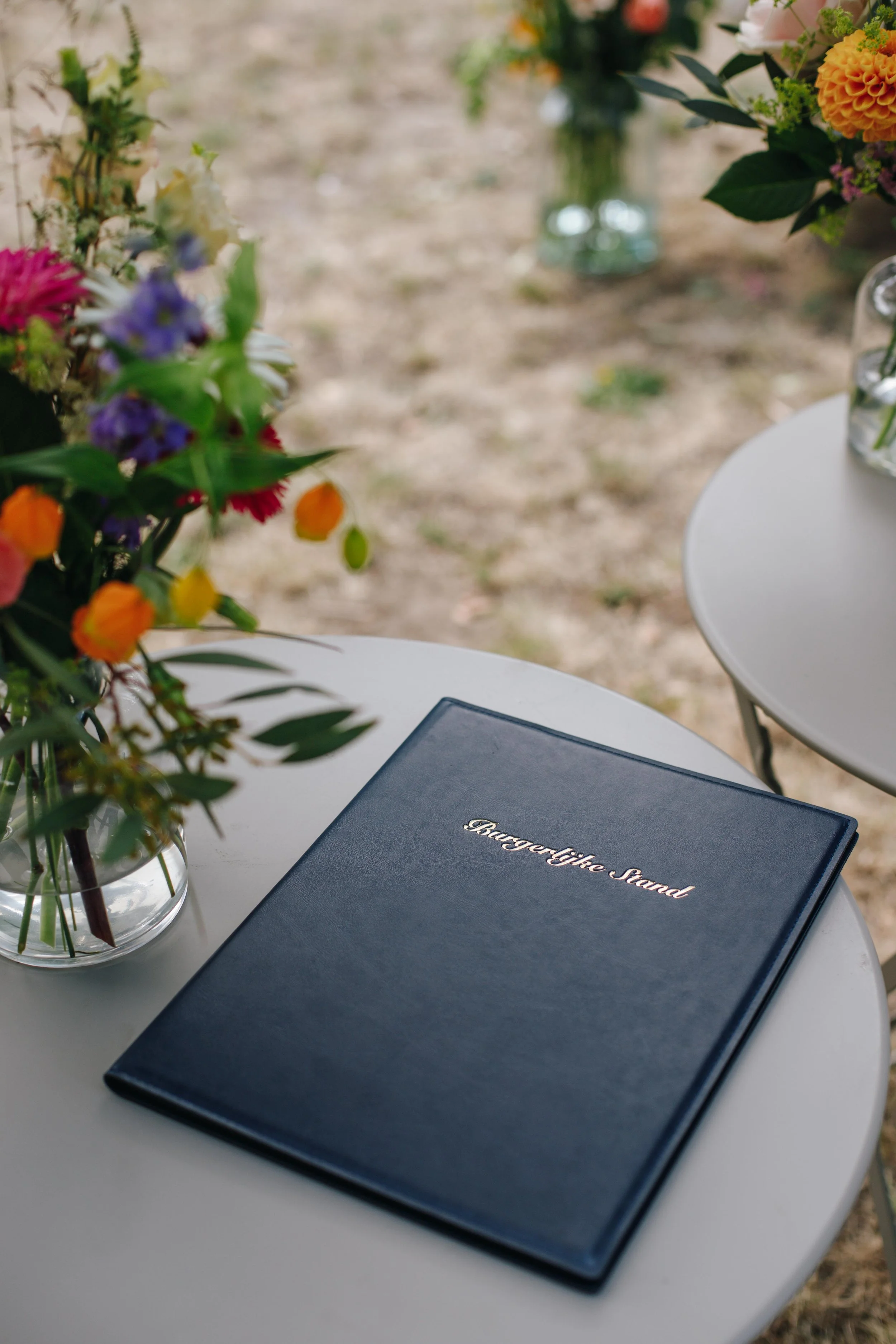 A white round table with a blue binder titled 'BurgerLife Stand' and a glass vase of colorful flowers outdoors.