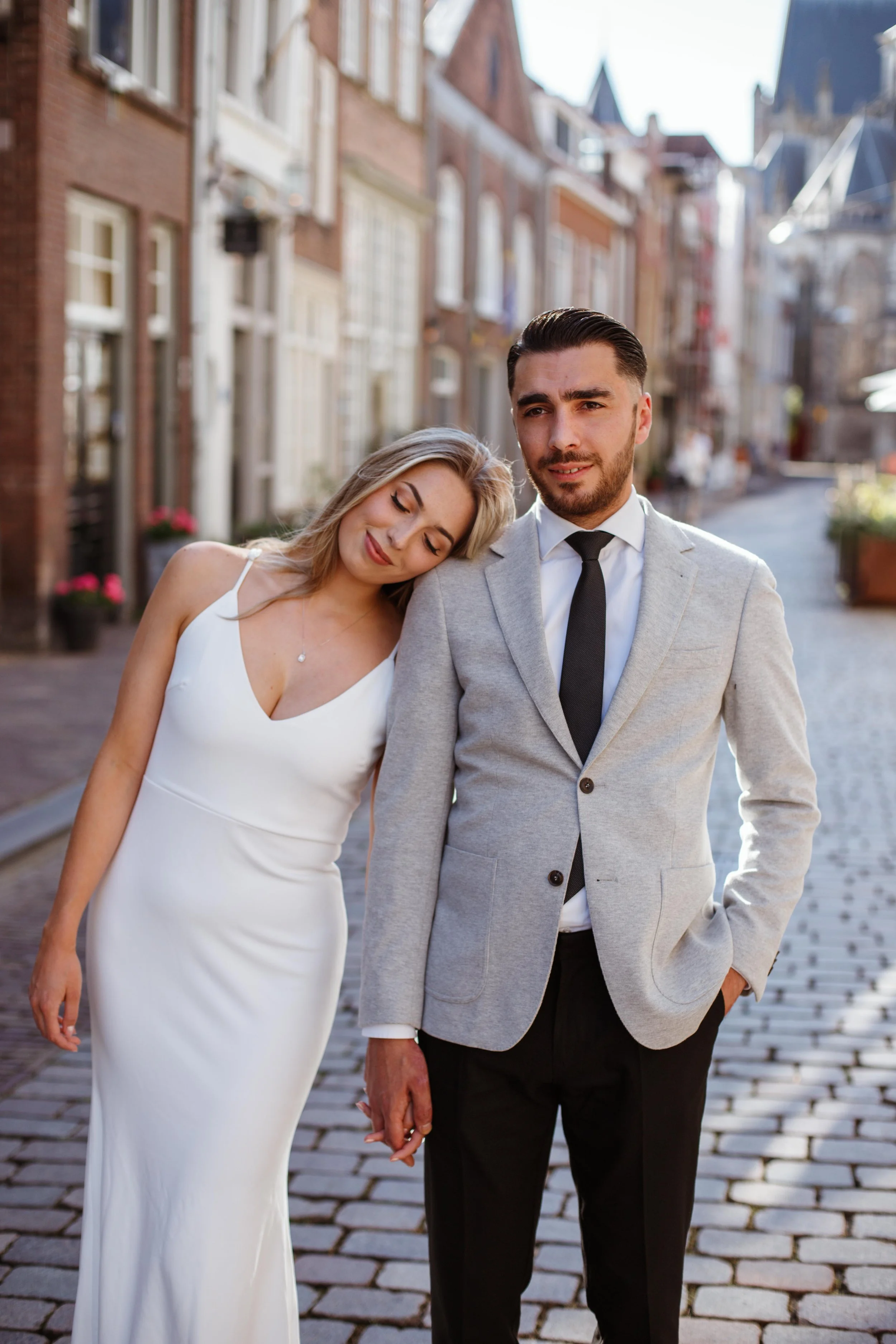 A man and woman holding hands on a cobblestone street in a European city, with buildings and flower pots in the background. The woman is wearing a white dress with her head leaning on the man's shoulder, who is dressed in a light gray blazer, white s