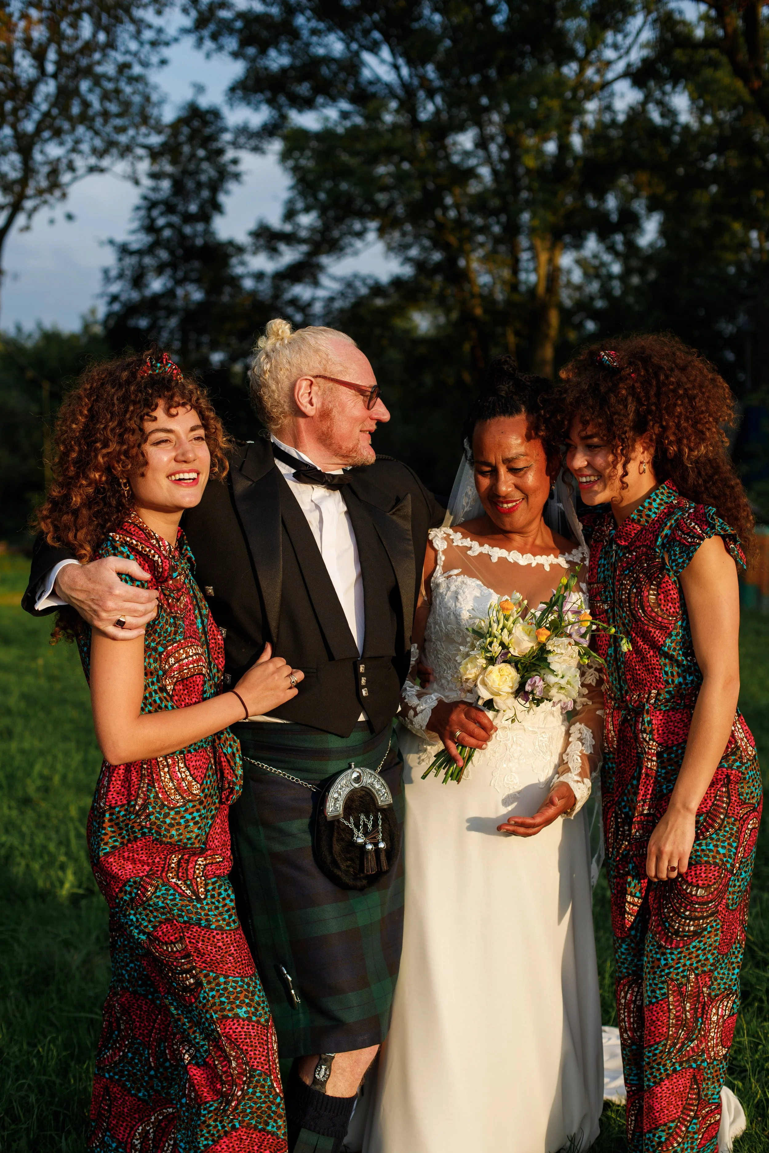 Four people, two women and one man, all smiling and embracing, standing outdoors during sunset. One woman is wearing a white wedding dress holding a bouquet, and the two other women are in colorful patterned dresses. One man is wearing a traditional 