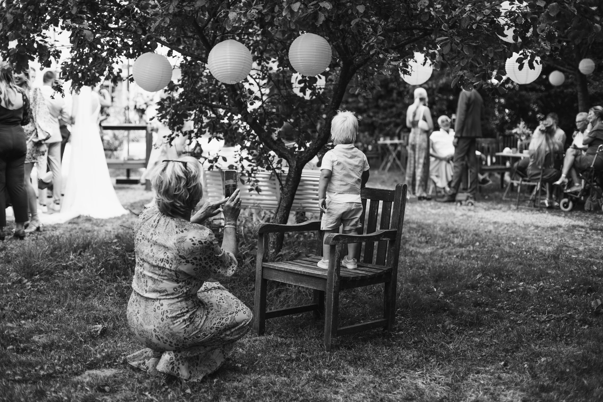 A young boy standing on a wooden chair under a tree with hanging paper lanterns, with an older woman crouching and taking a photo of him using a smartphone at an outdoor gathering, surrounded by other people socializing and sitting at tables.