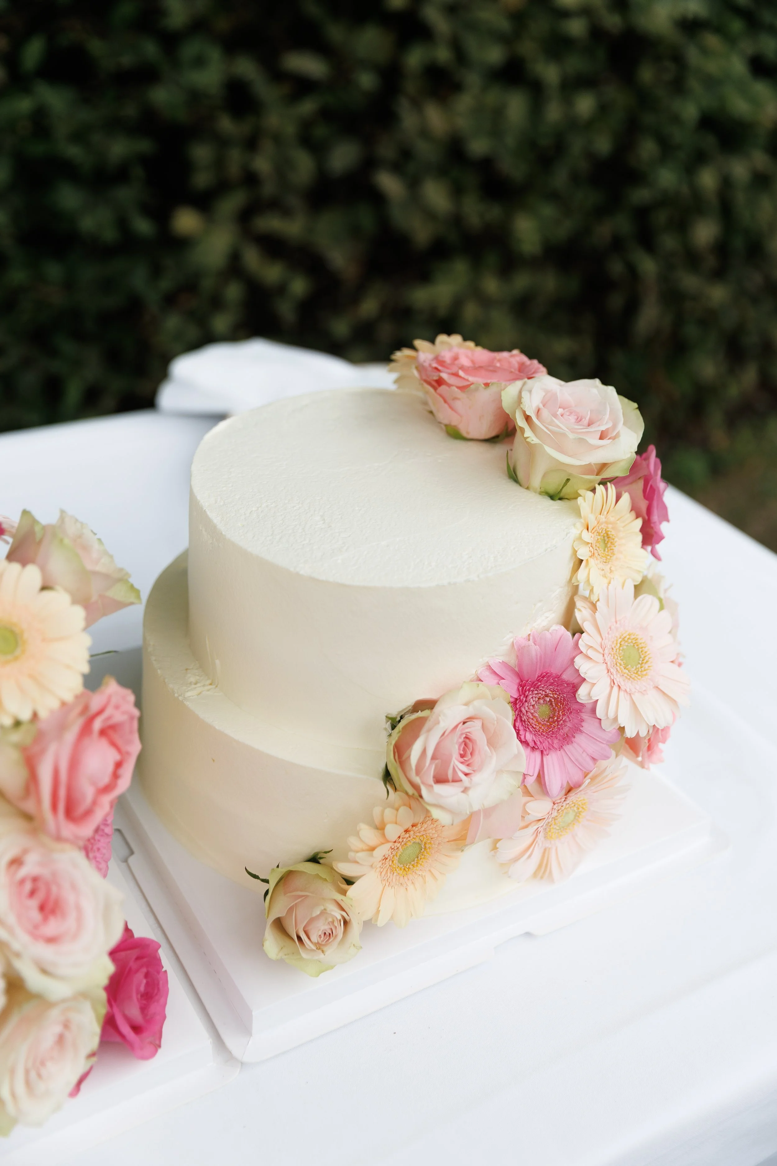 A two-tier white wedding cake decorated with pink and cream roses and pink daisies, placed on a white tablecloth.