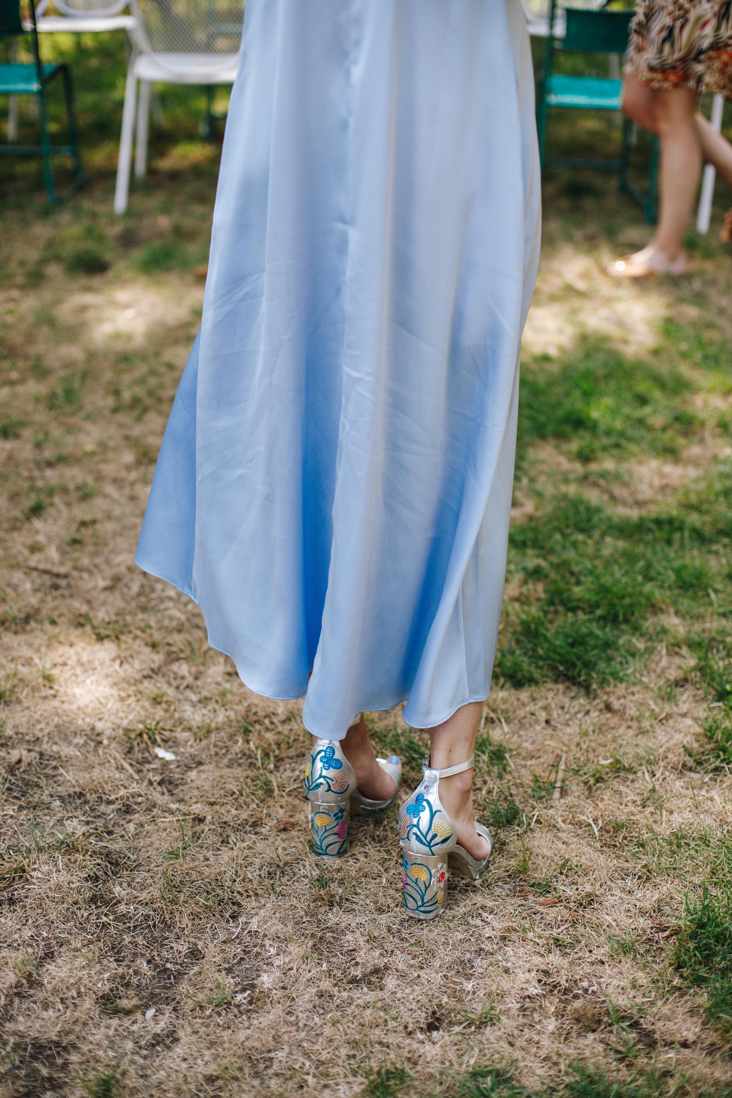Woman wearing a light blue dress and colorful high-heeled sandals standing outdoors on grass, with chairs visible in the background.