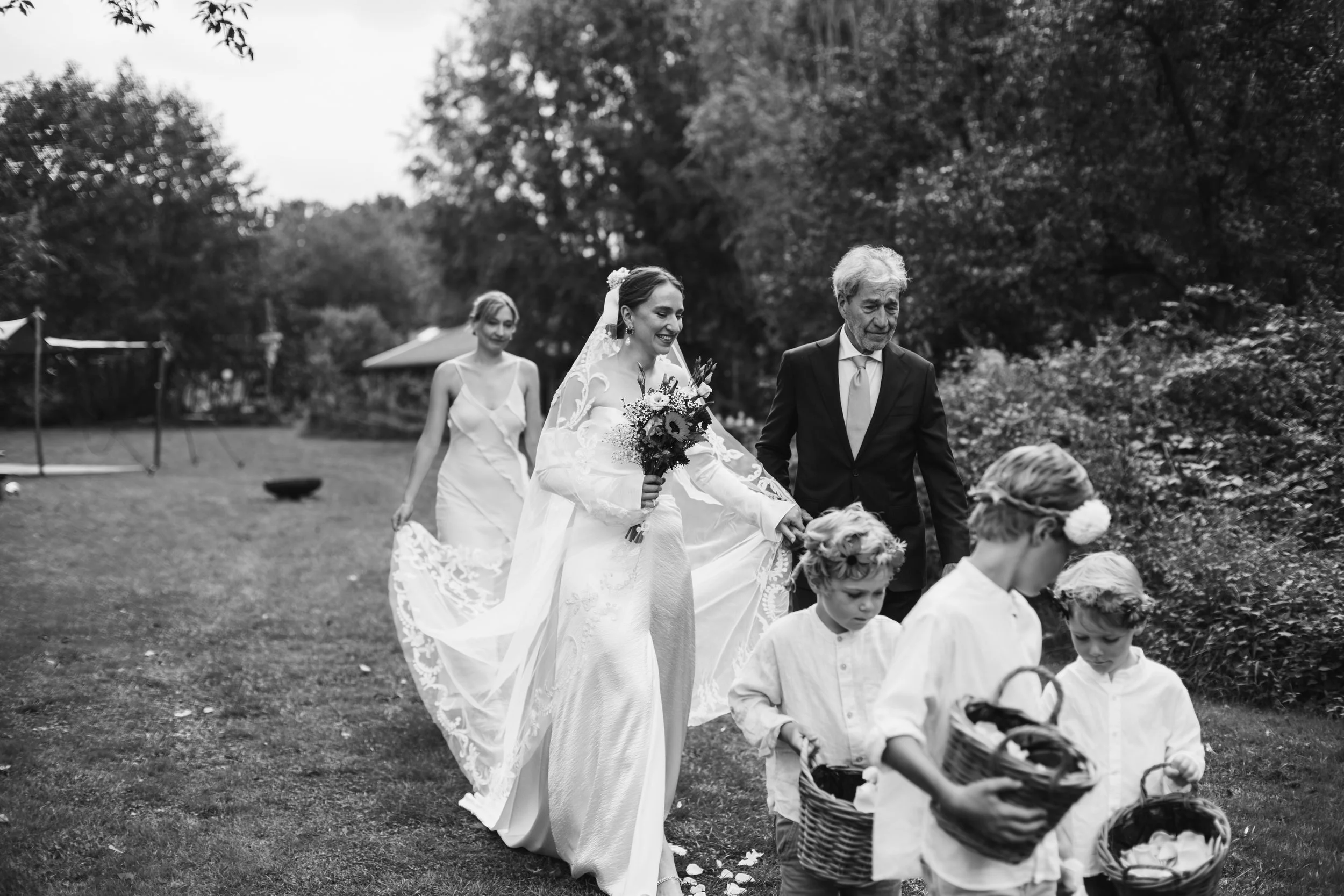 Black and white photo of a bride walking outdoors with a man, accompanied by three young boys carrying baskets, with a woman following behind, all surrounded by trees.