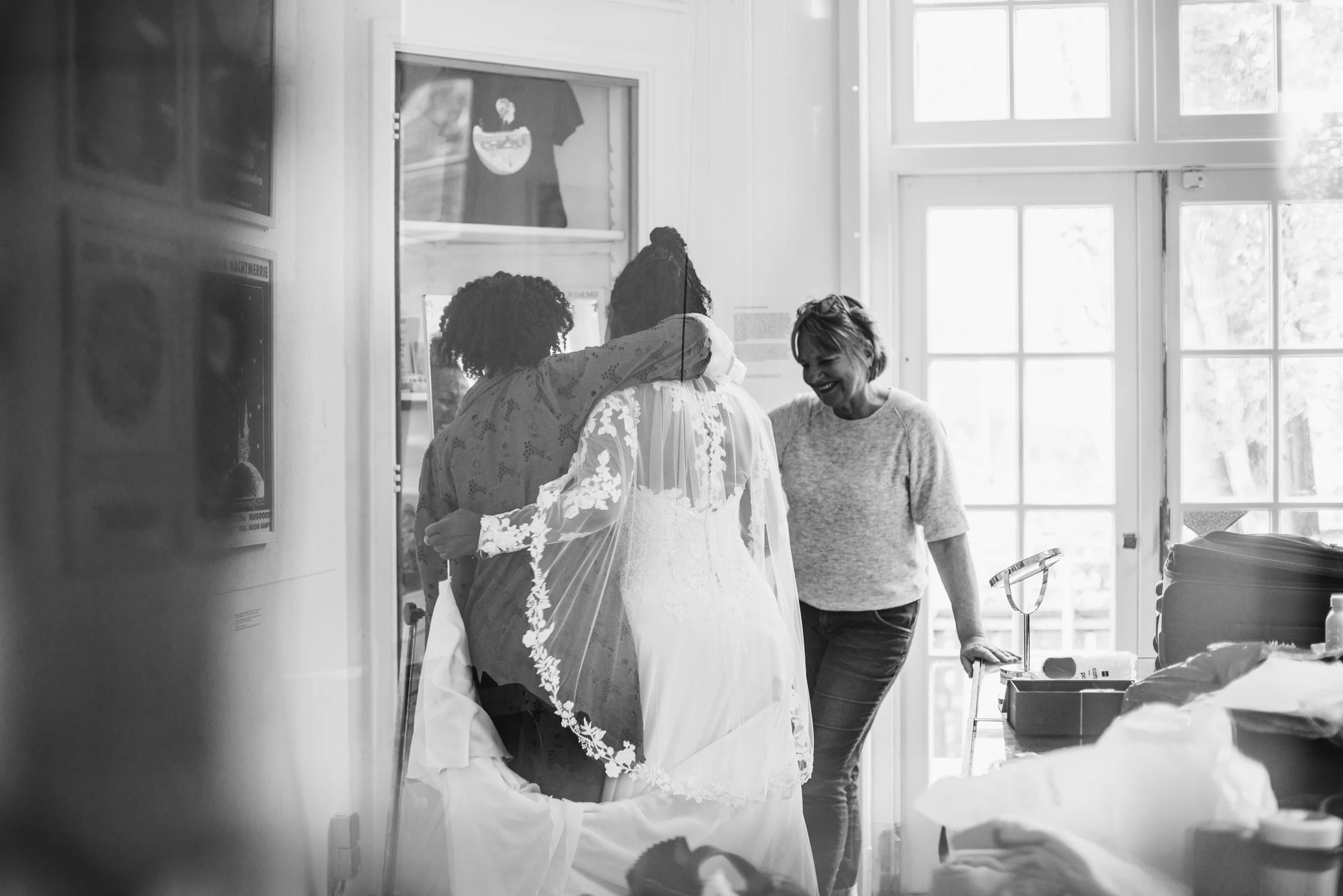 Women preparing for a wedding, with the bride in a wedding dress and veil, in a brightly lit room with large windows.