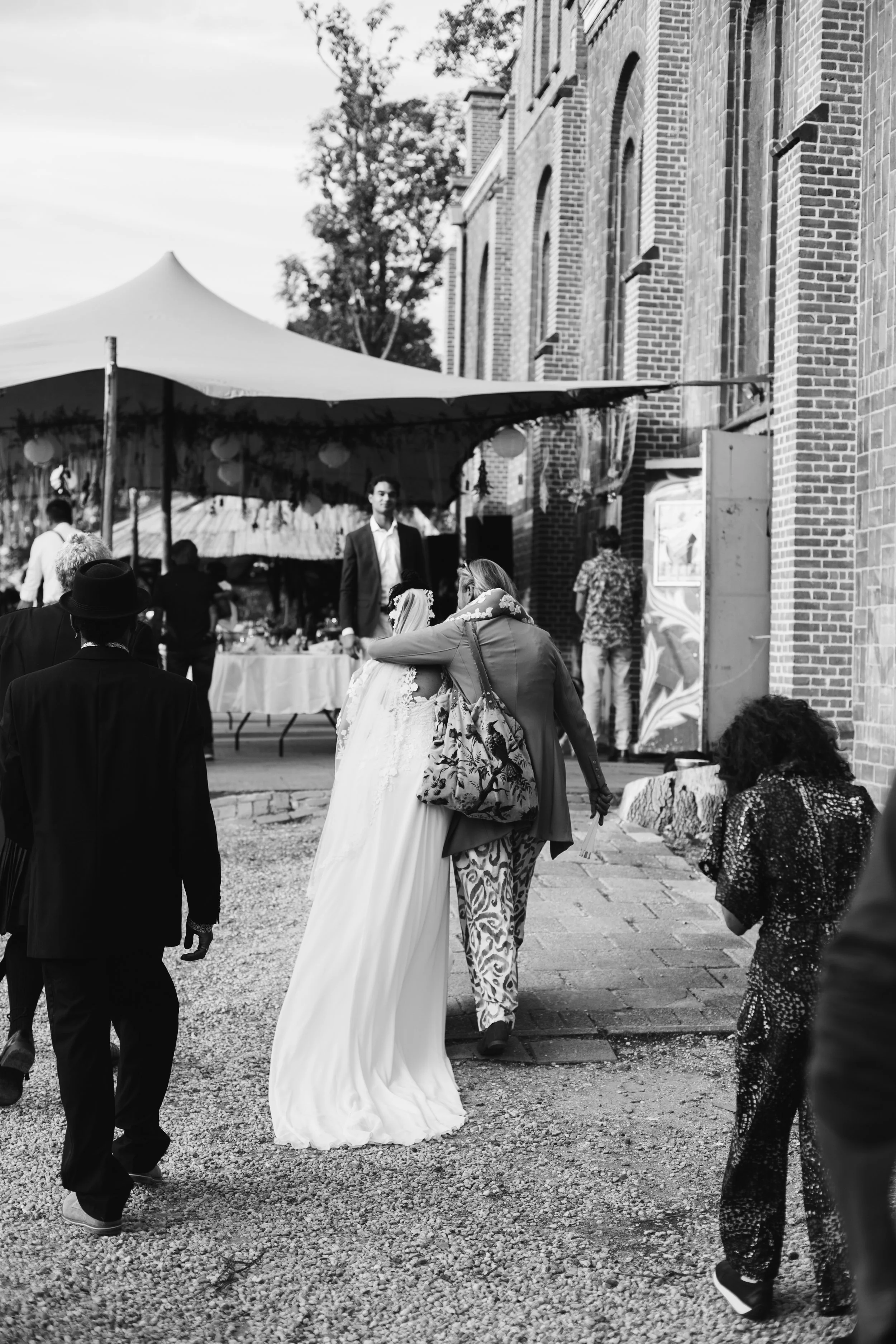 A bride in a wedding gown hugging a woman with a patterned bag outside a brick building, with a tent and people in the background.