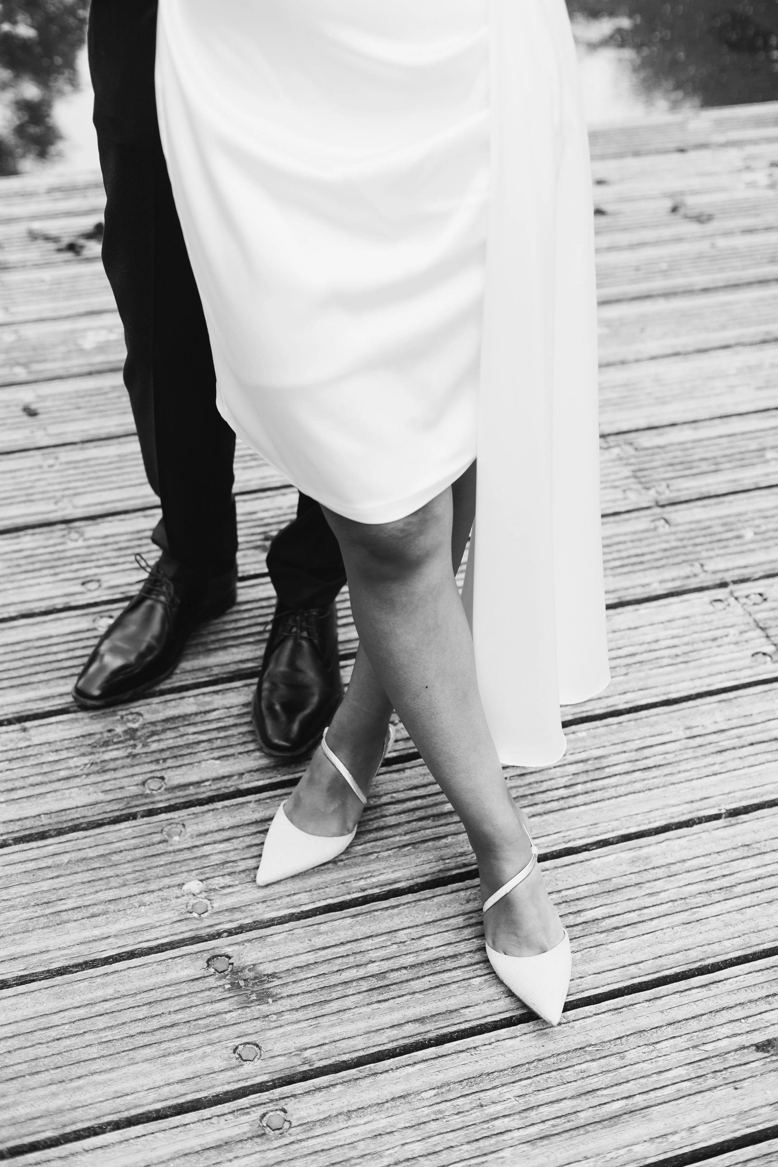 Close-up of a couple's legs and feet on a wooden pier, with the woman in white high heels and the man in black dress shoes, both dressed formally.